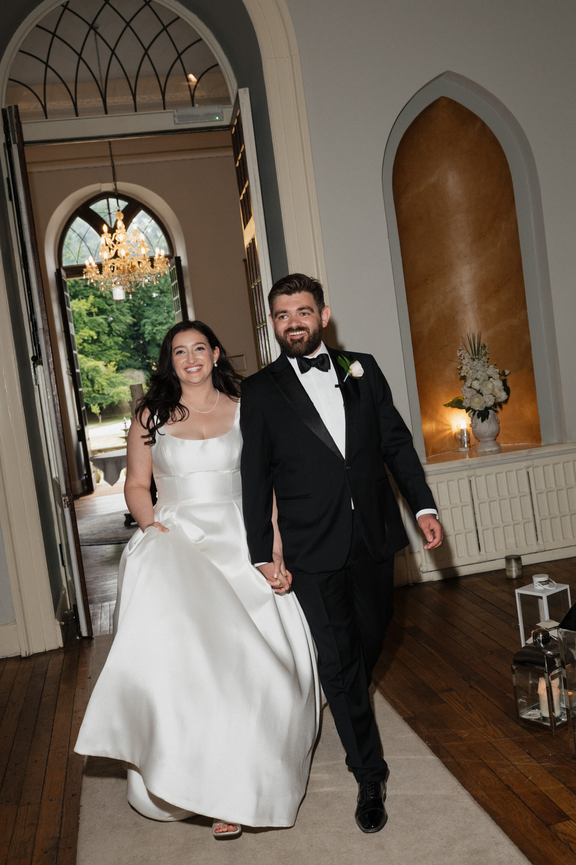 A bride in a white dress and a groom in a black tux walk inside, holding hands and smiling, with greenery and a chandelier in the background.