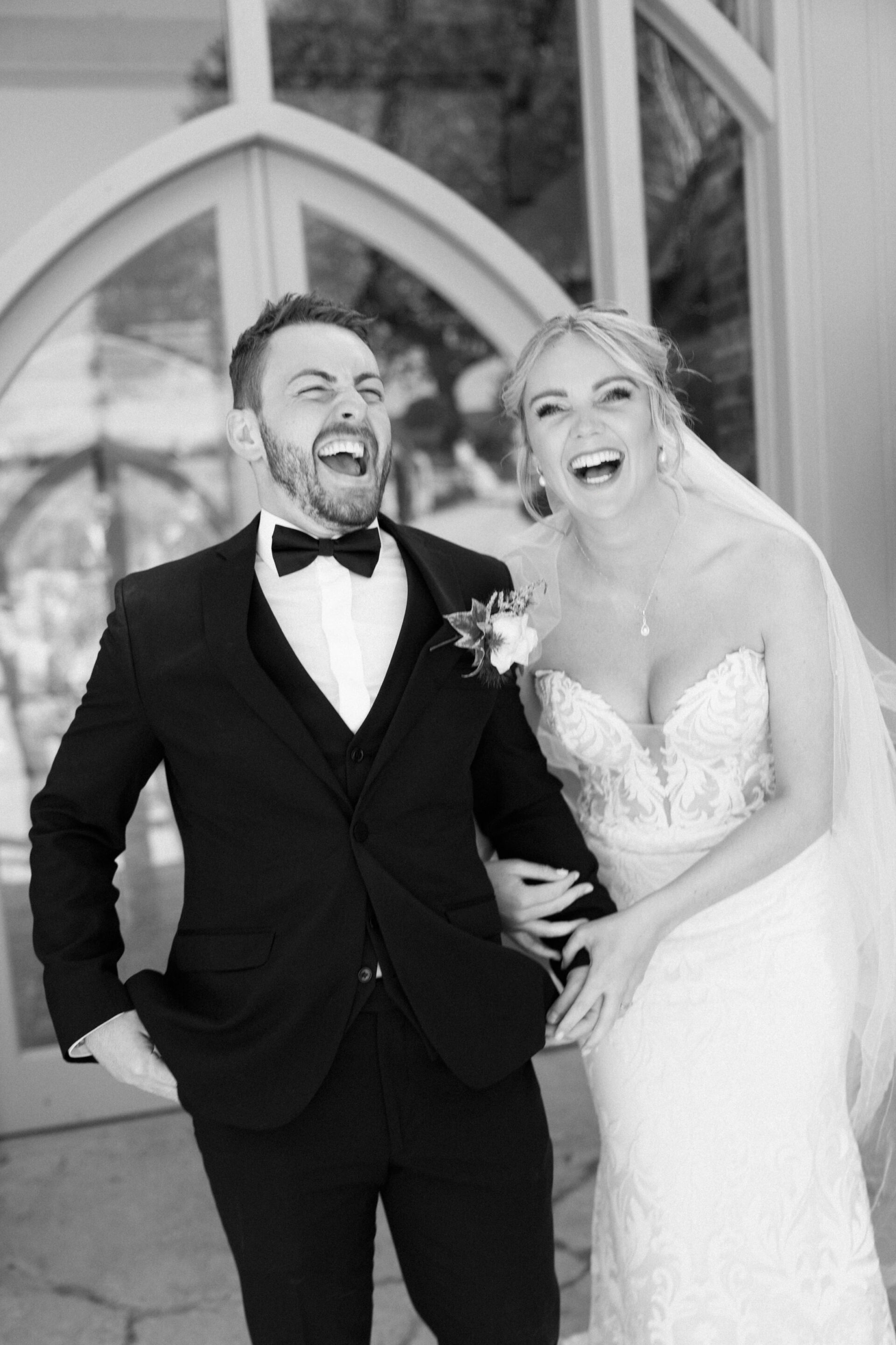 A bride and groom dressed in their wedding clothes are standing outside, holding hands and laughing together.