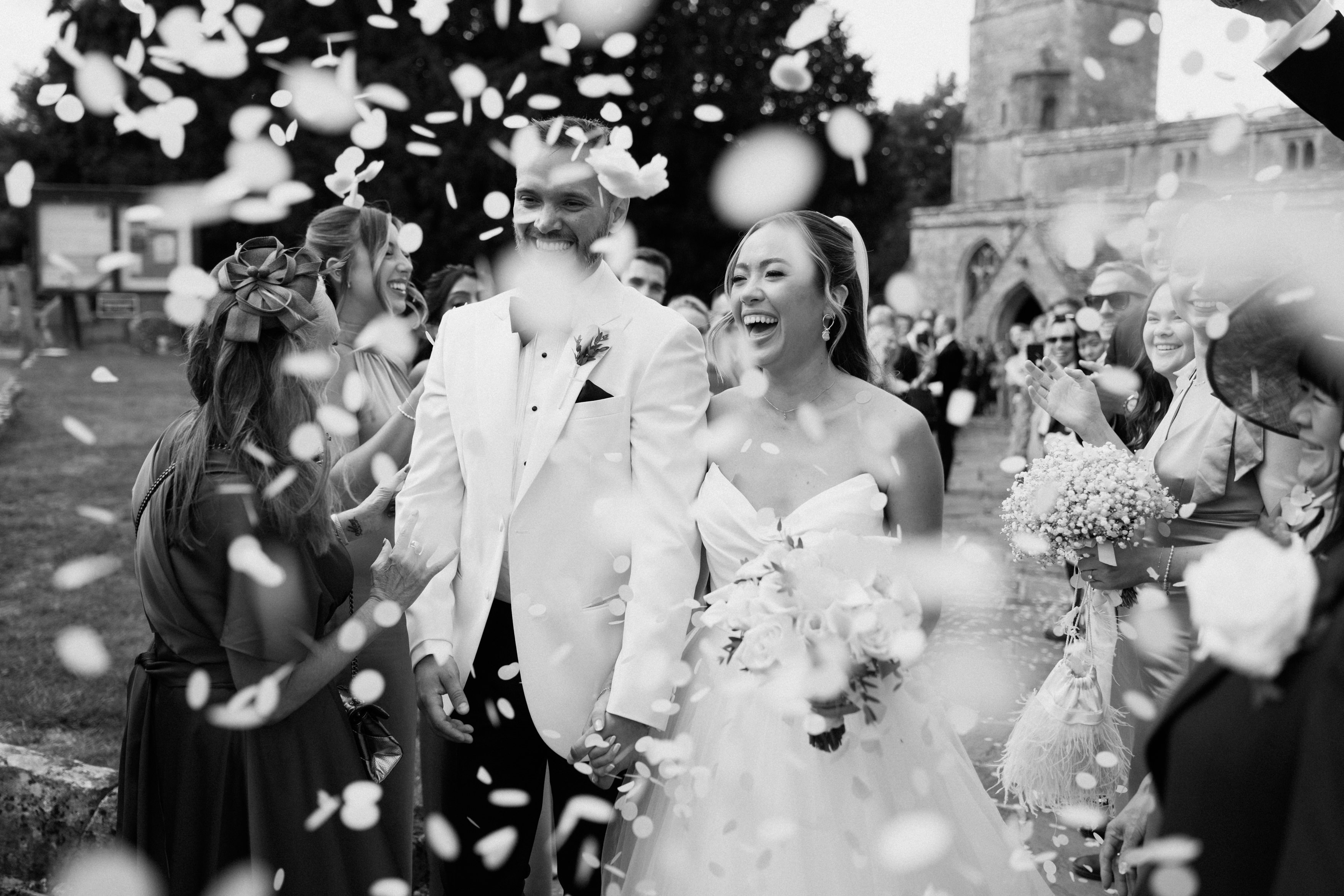 The bride and groom walk together holding hands while guests toss flower petals at them outside near a stone building.
