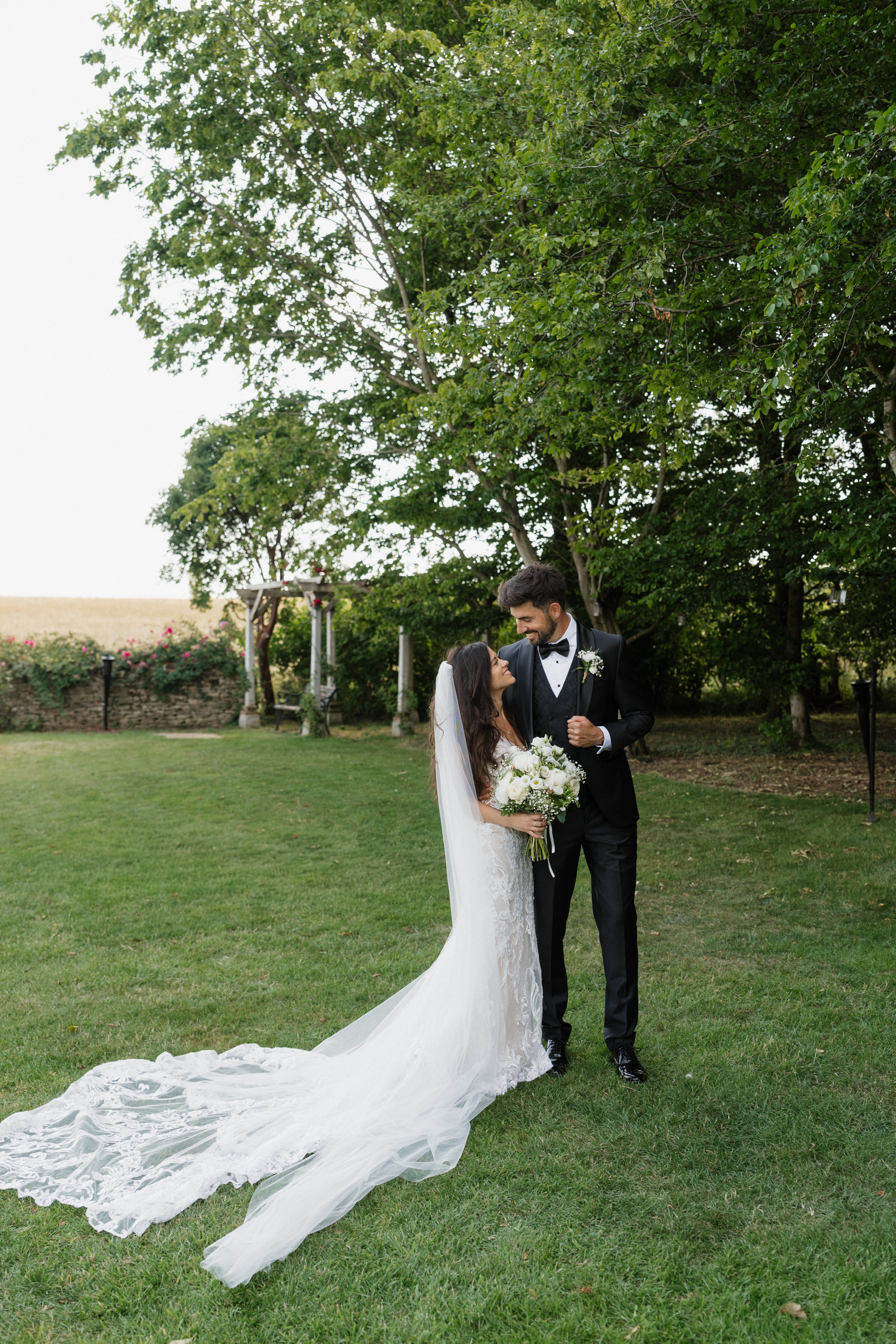 A bride wearing a white dress and veil is standing on the grass, holding some flowers and looking at the groom, who's dressed in a black suit. There are trees and green plants behind them.