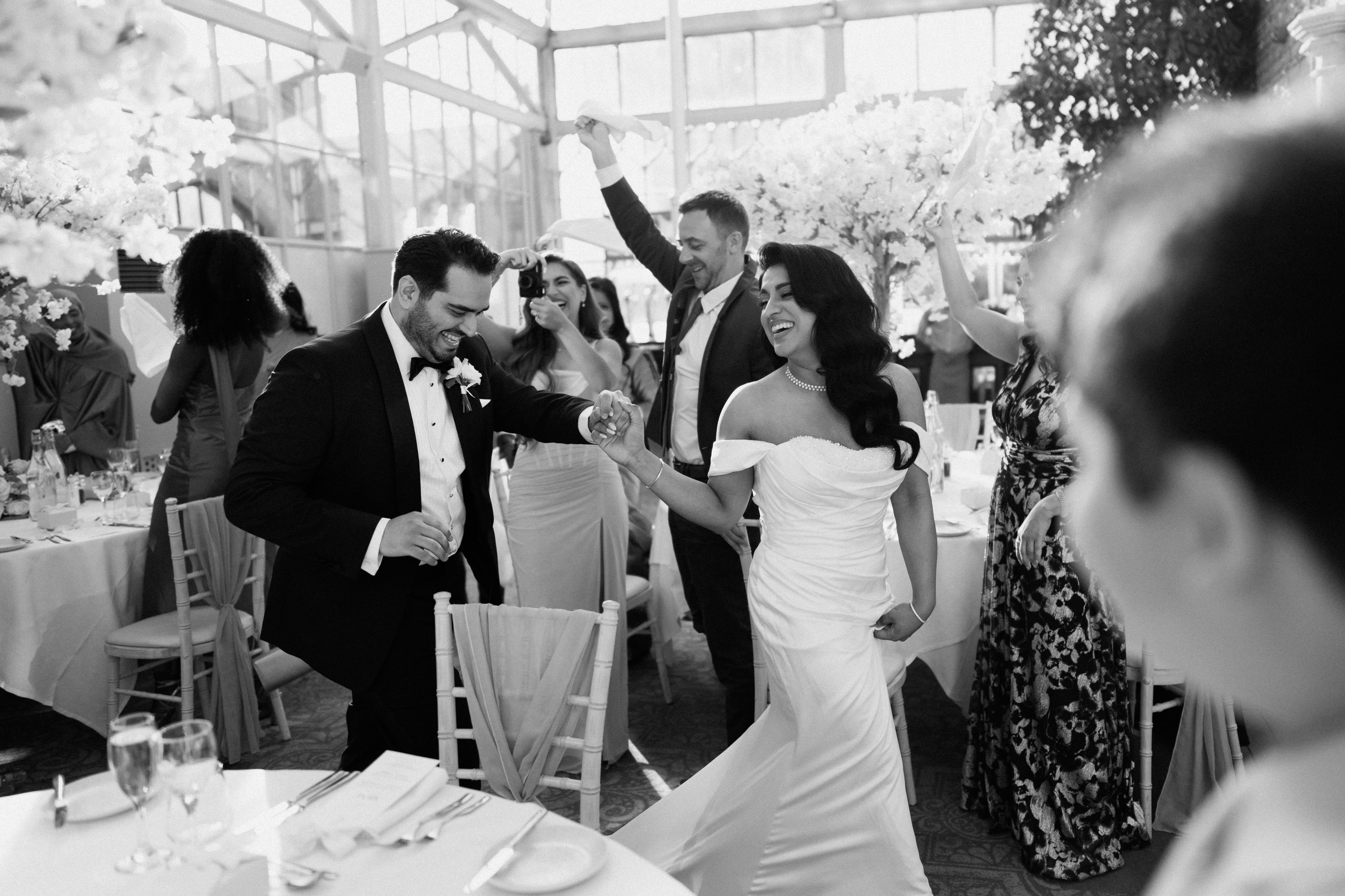 The bride and groom hold hands and smile as guests cheer for them at their wedding in a brightly decorated room.