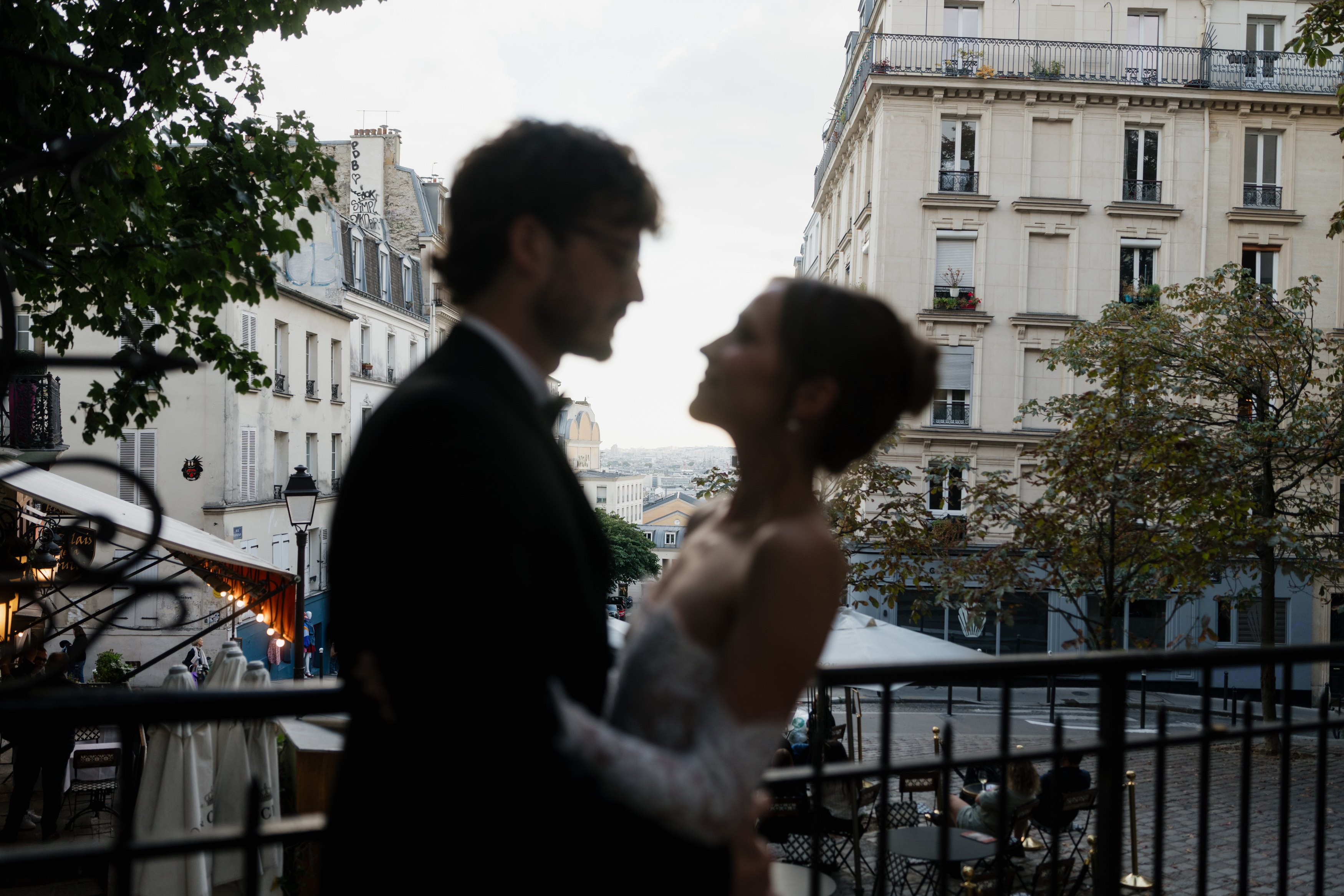 A couple stands side by side on a balcony, with out-of-focus Paris buildings and trees visible behind them.