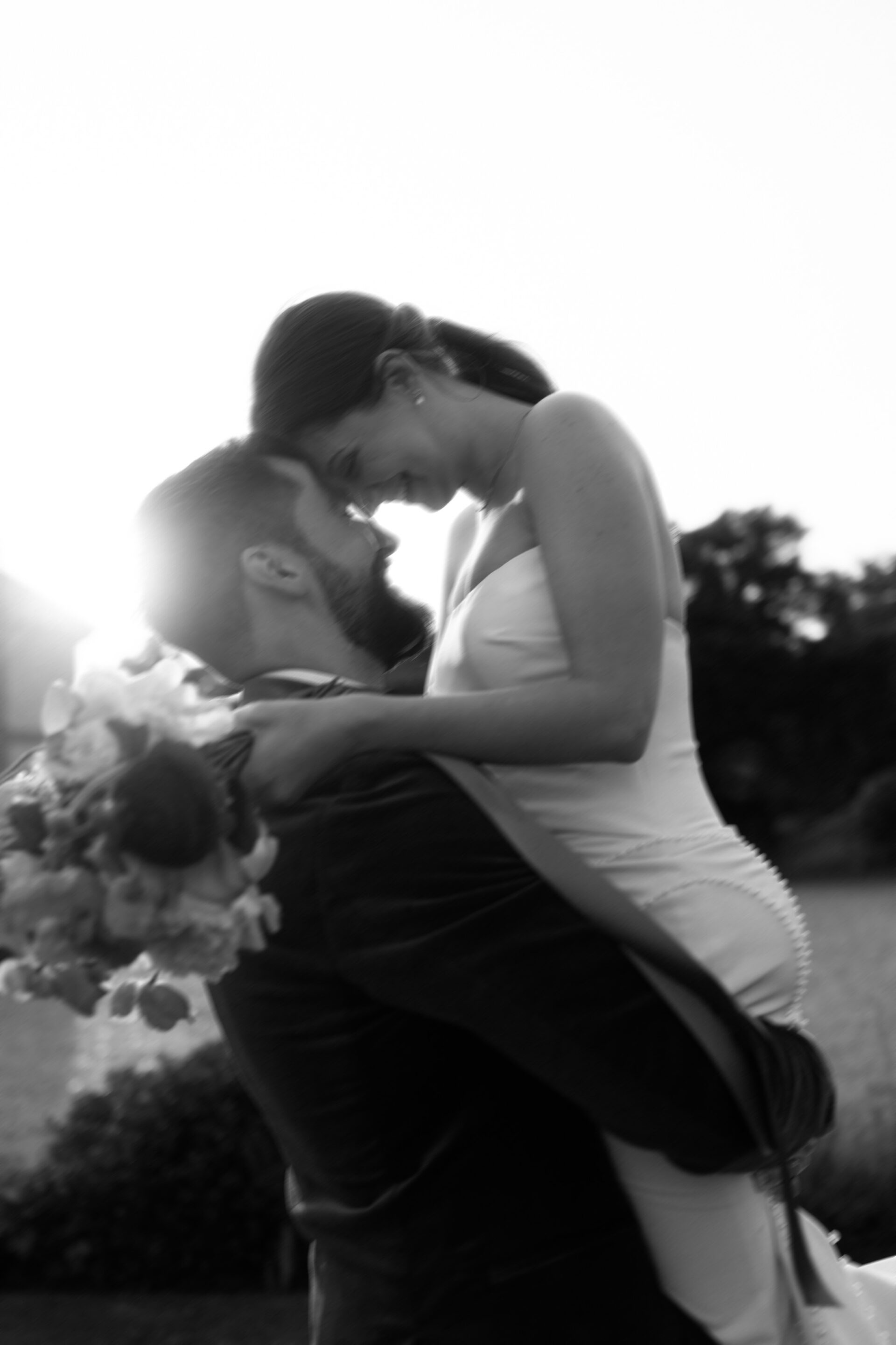 A groom picks up his bride outside at sunset. She’s holding a bouquet, and they’re smiling with their foreheads touching. The photo is in black and white.