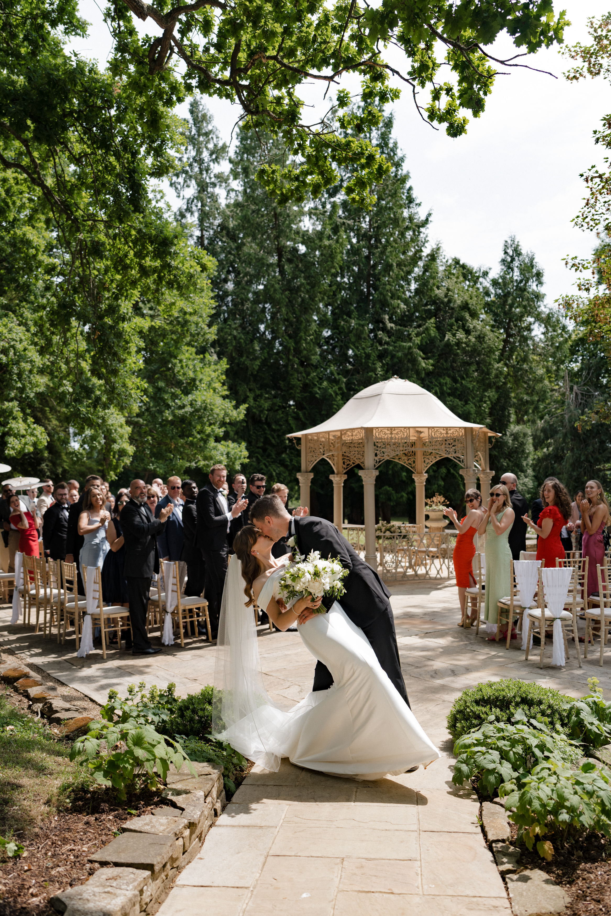 A newly married couple kisses on a stone path outside while their guests clap for them. There’s a wooden gazebo and lots of greenery behind them.