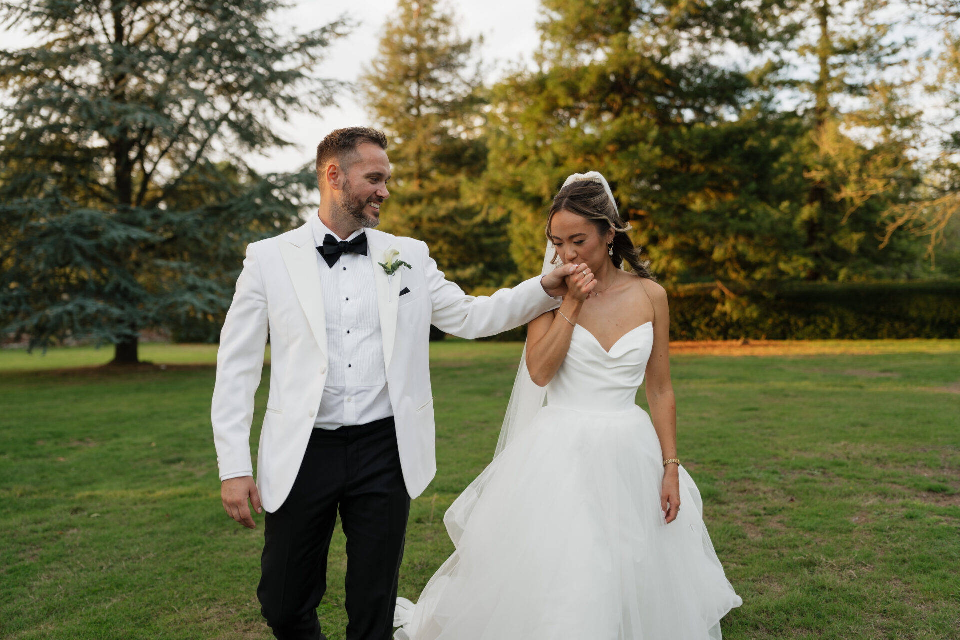 A groom in a white tux kisses his bride’s hand as they walk on grass, with trees behind them. She’s wearing a strapless white dress.
