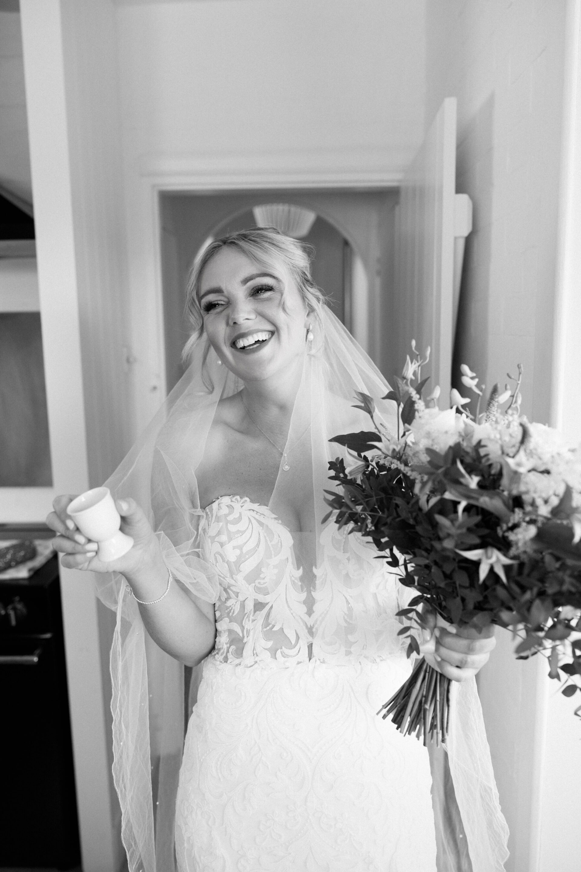 A bride in a white dress and veil smiles inside, holding a bouquet and a small object in her hand.