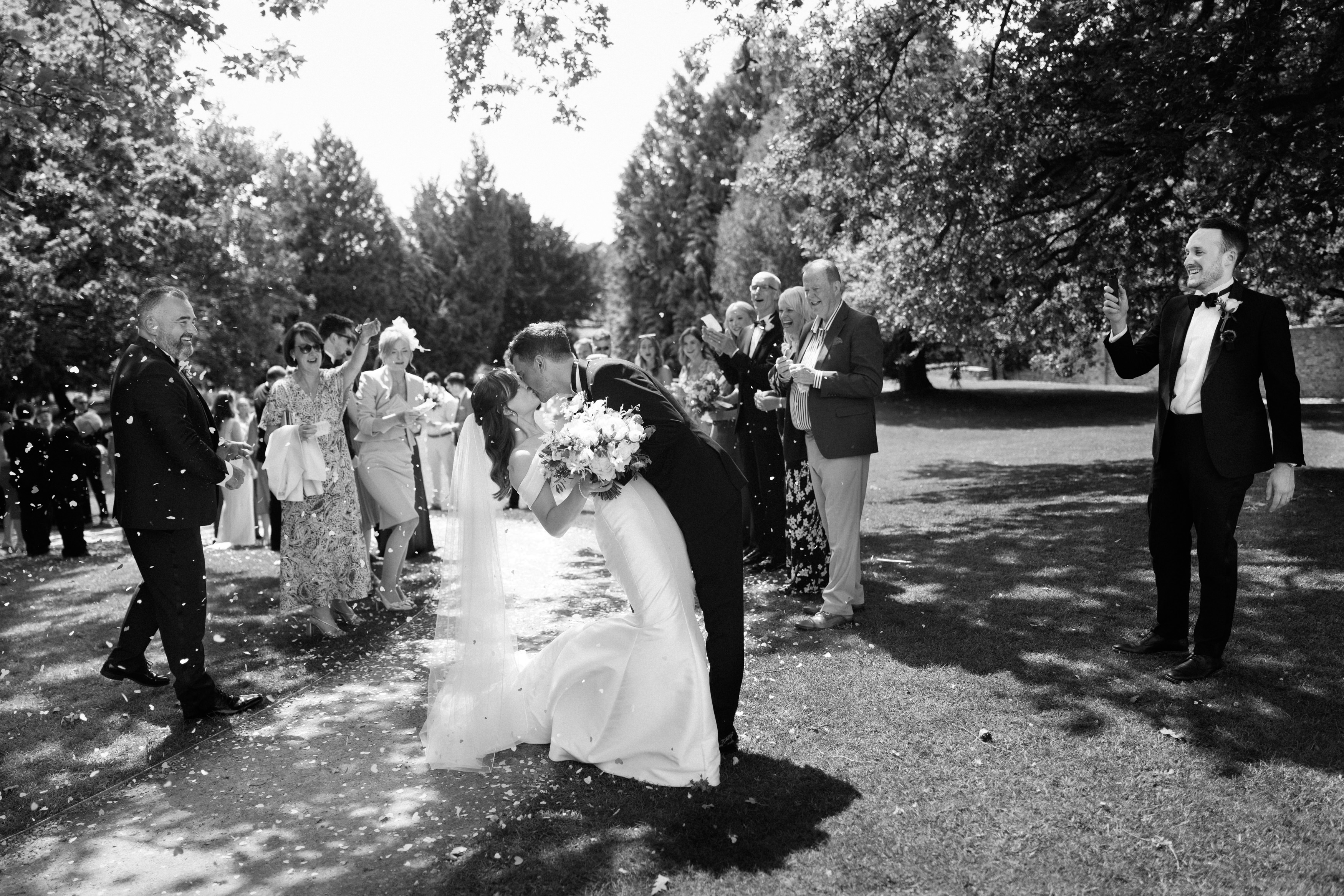 A bride and groom kiss outside while their wedding guests stand nearby. Some people are tossing confetti, and everyone is under the trees on a sunny day.