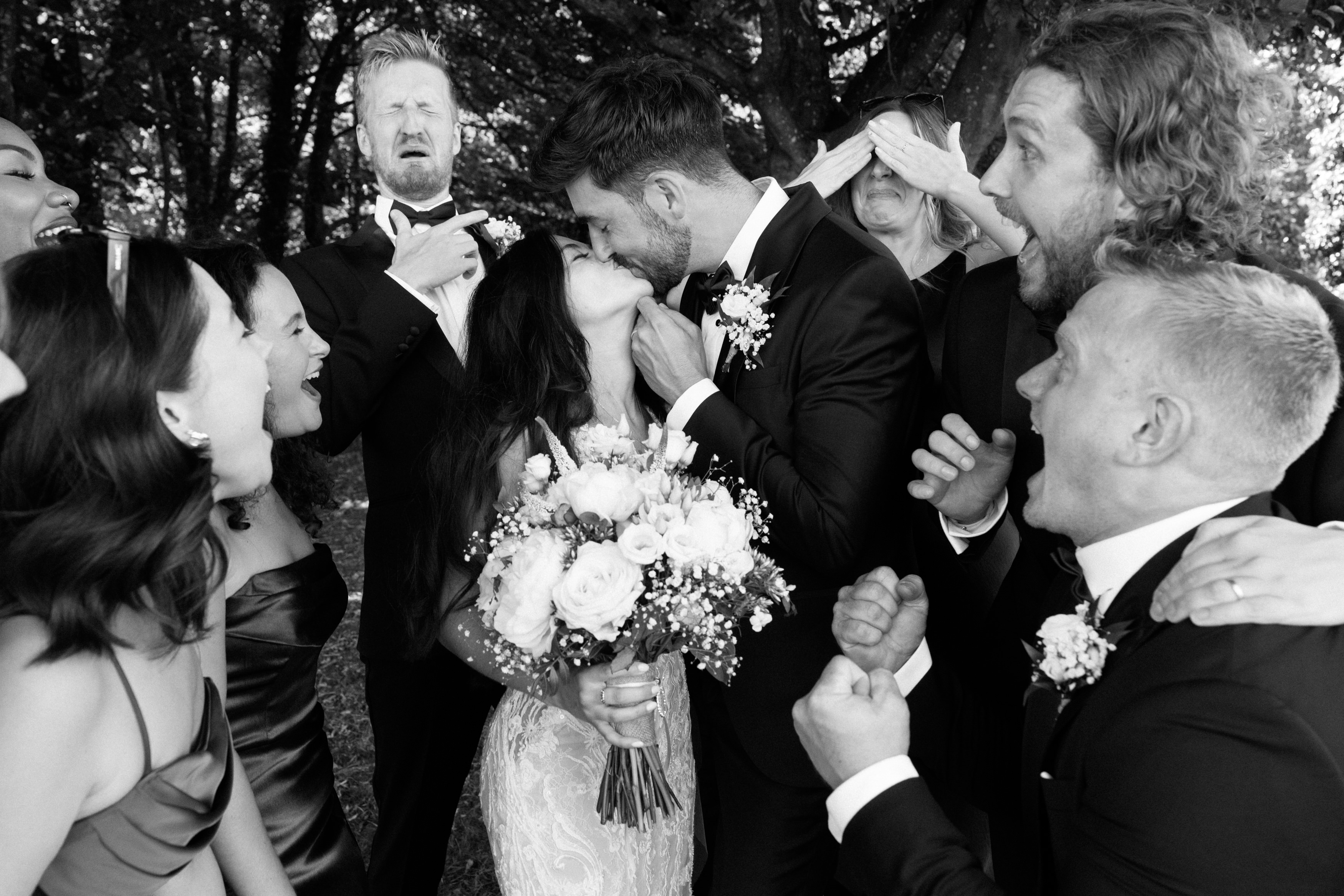 A bride and groom share a kiss outside while their wedding guests, dressed up, cheer and laugh around them.