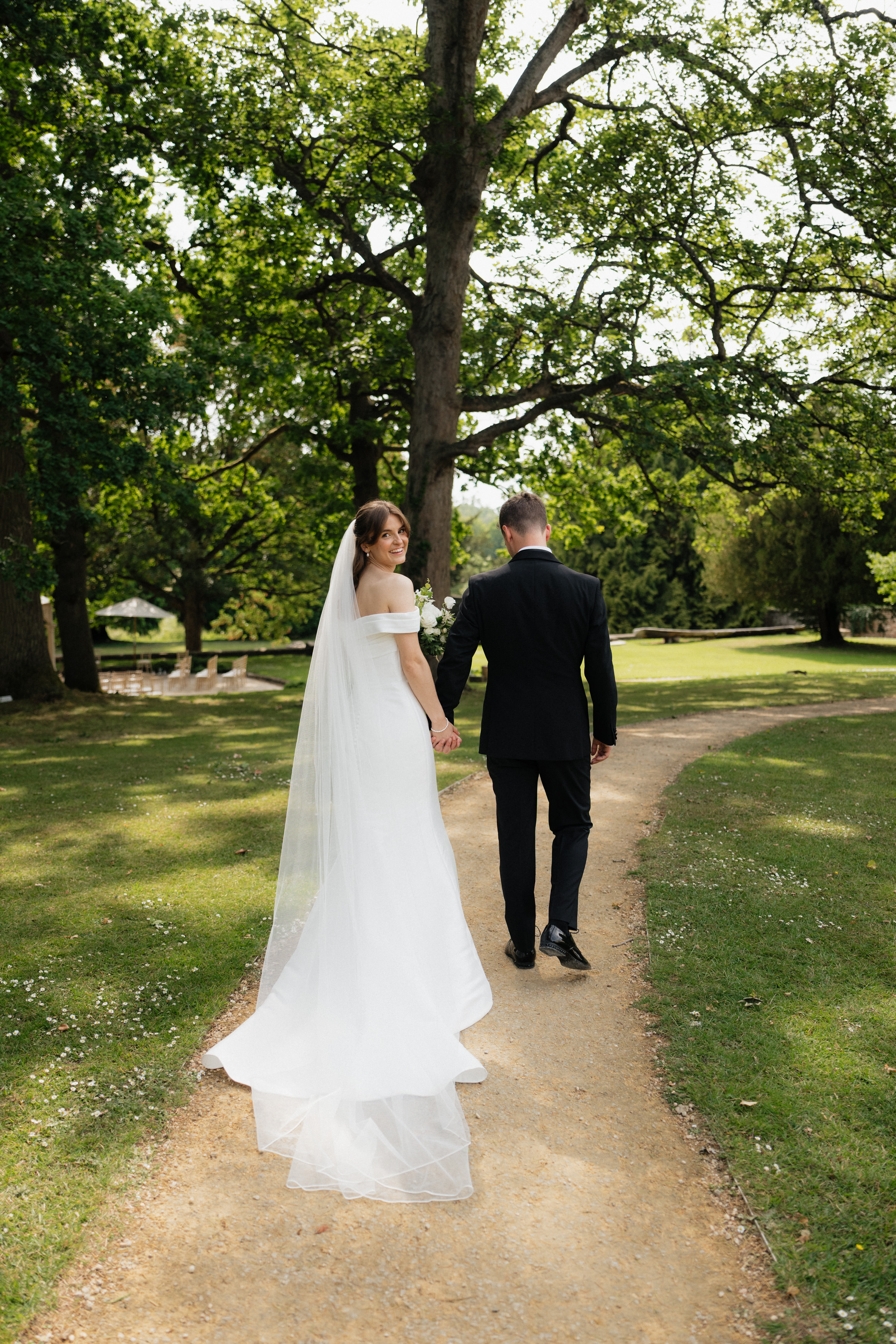 A bride wearing a white dress and veil is holding hands with the groom, who’s dressed in a black suit. They’re walking together along a garden path with trees and plants all around them.