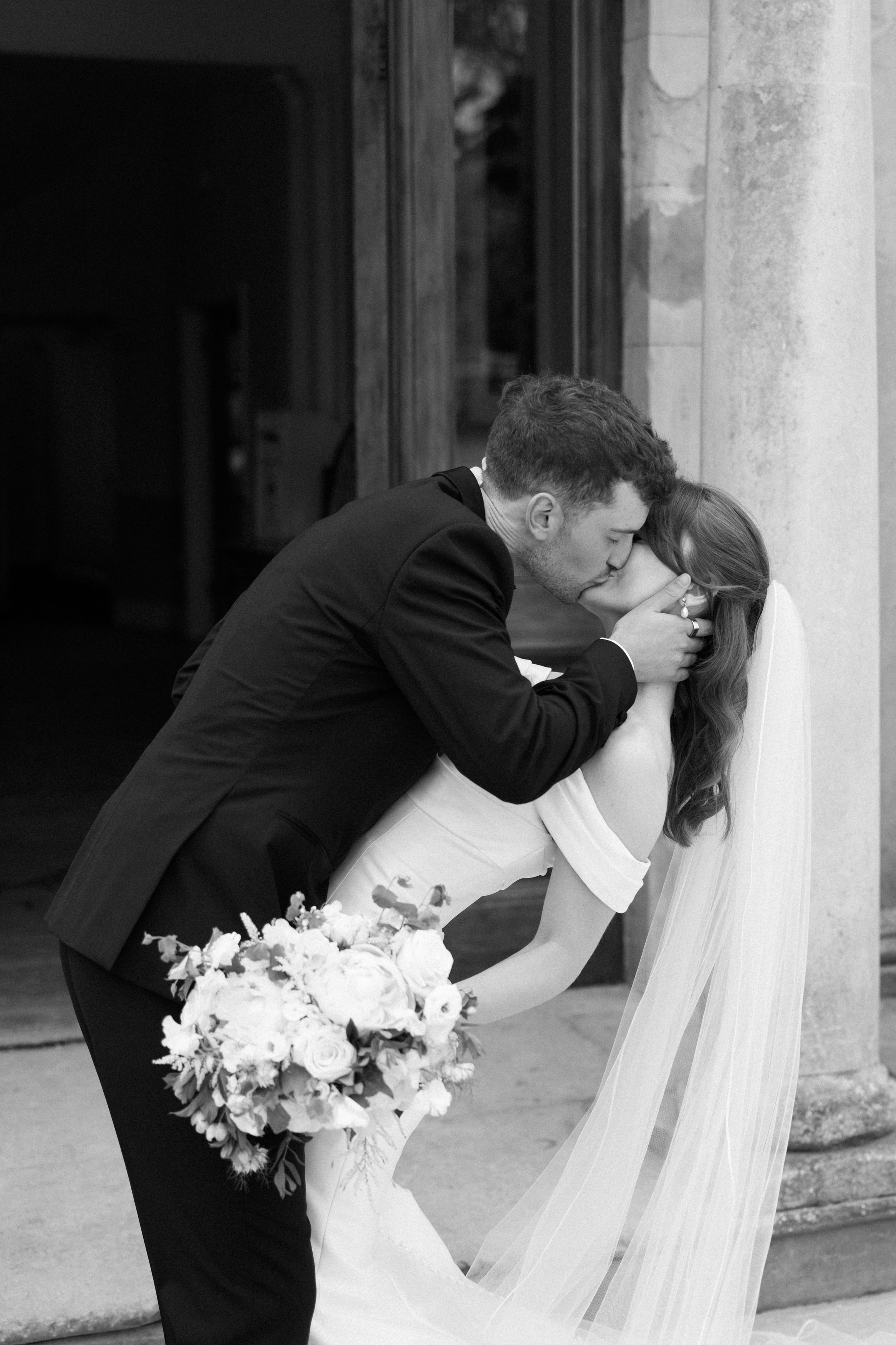 A man in a suit kisses his bride, who’s wearing a white dress and veil, as he dips her and she holds a bouquet of flowers.