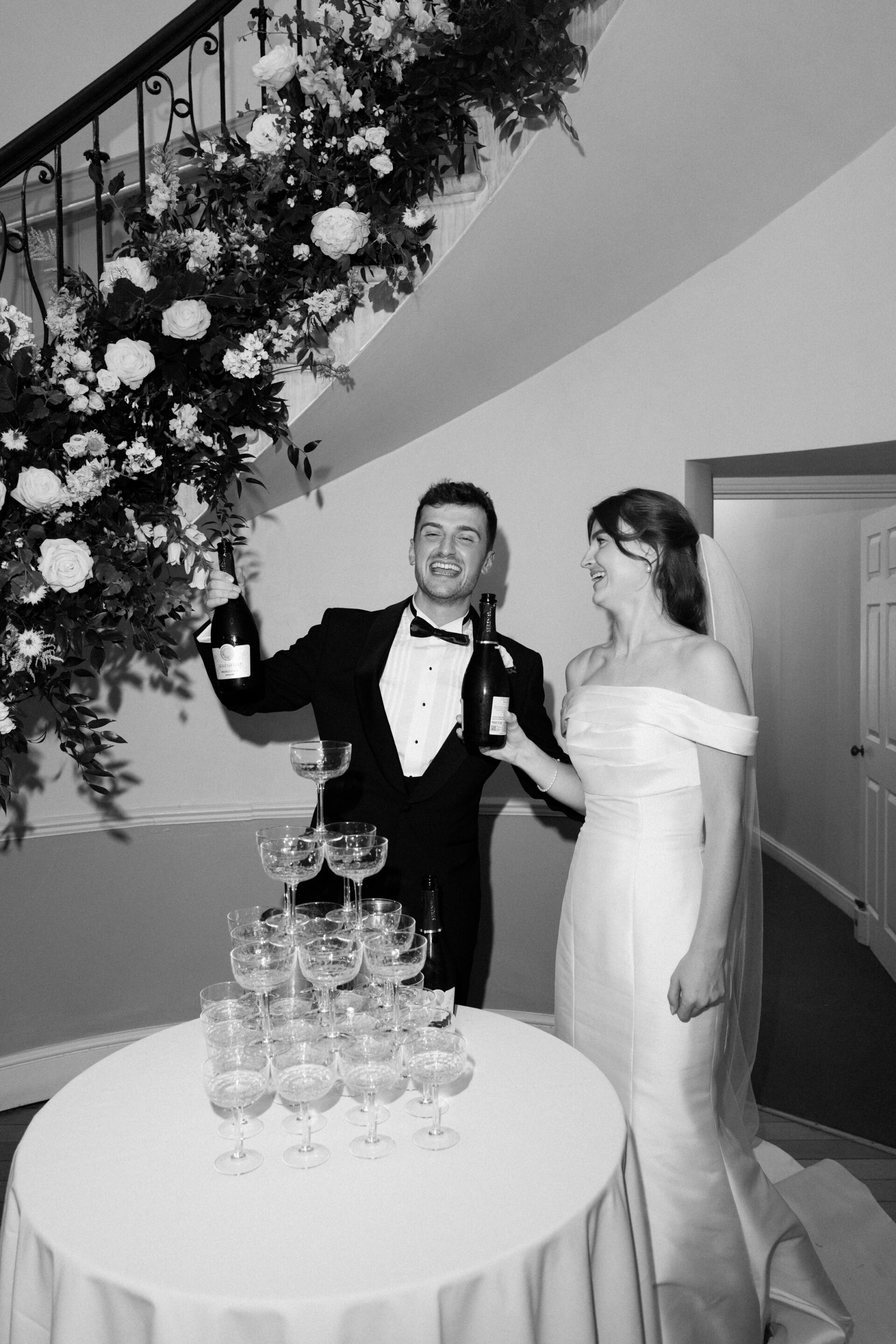A bride and groom stand next to a champagne tower, each holding a bottle of champagne, under some flowers by the stairs.