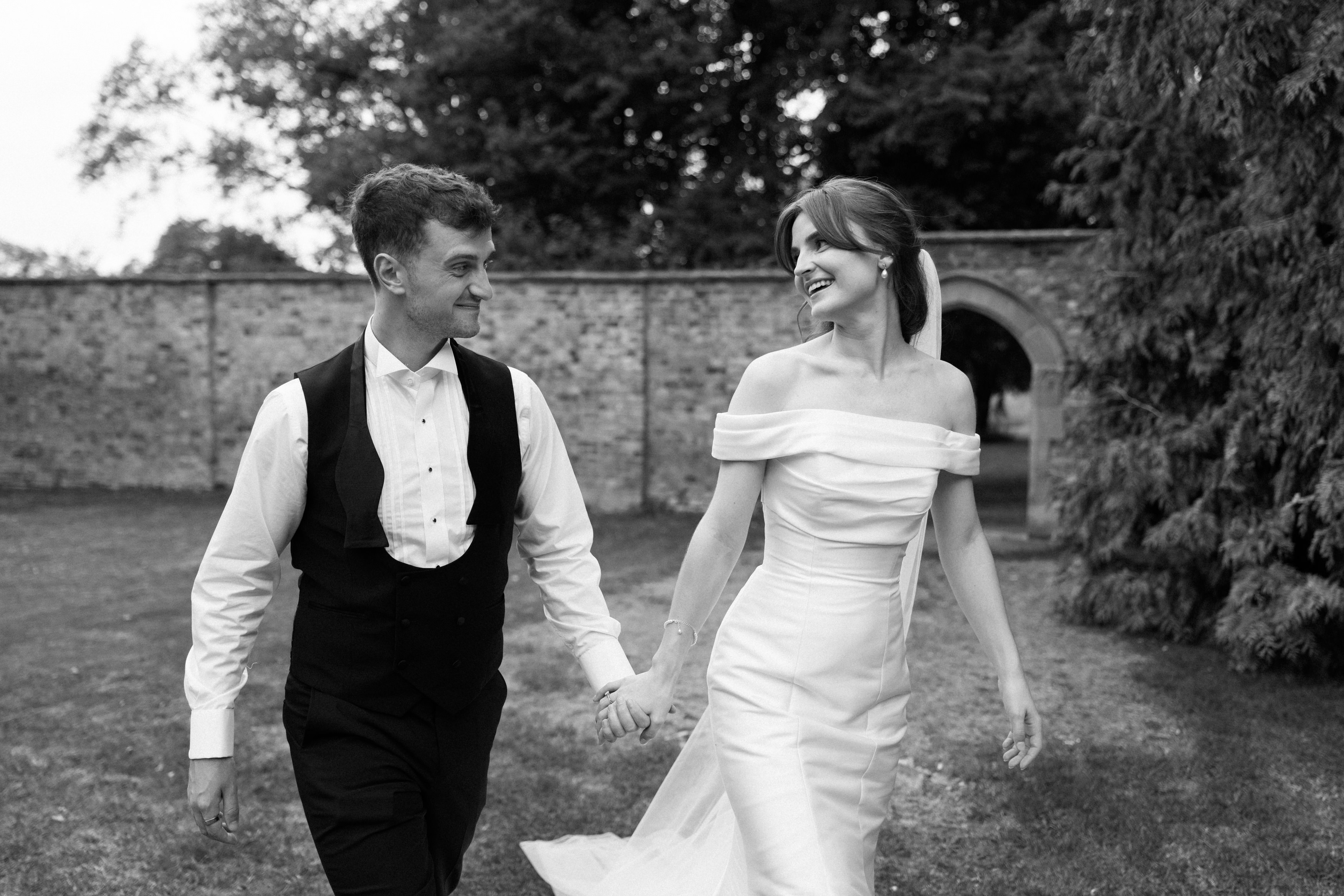 A couple dressed in wedding clothes are walking outside, holding hands and smiling at each other. There are trees and a brick wall behind them.
