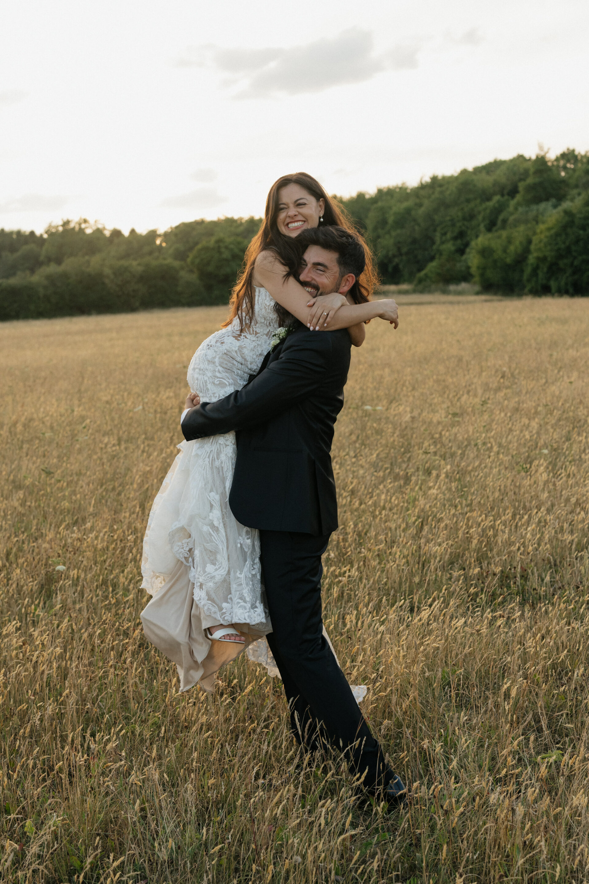 A man in a black suit hugs and lifts up a woman in a white dress while they both smile, standing in a field with trees behind them.