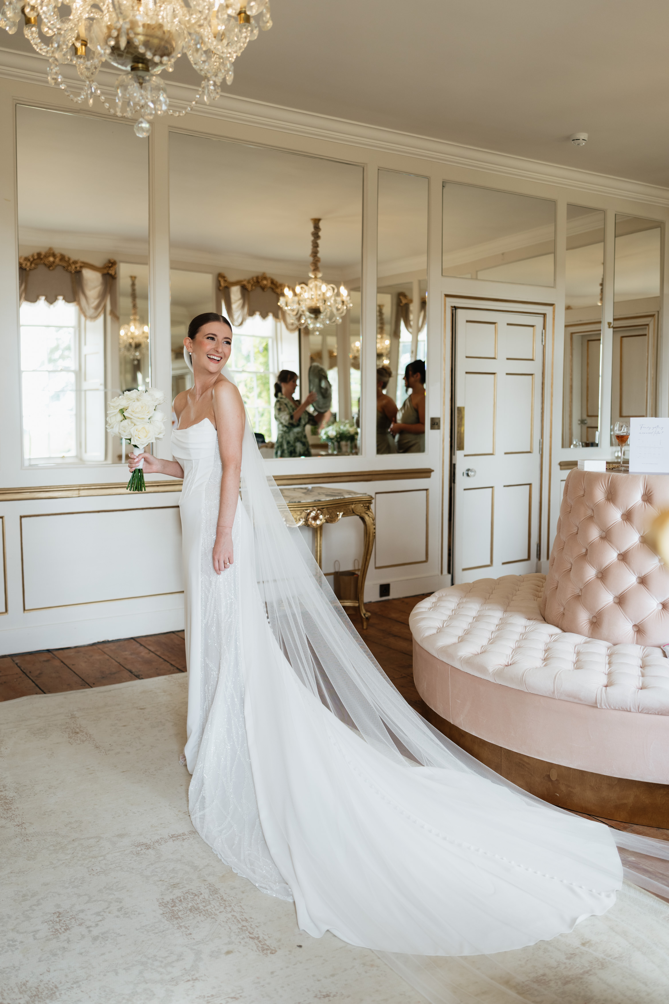 A bride wearing a white dress and veil smiles and holds flowers, standing in a fancy room with chandeliers and fancy decorations.