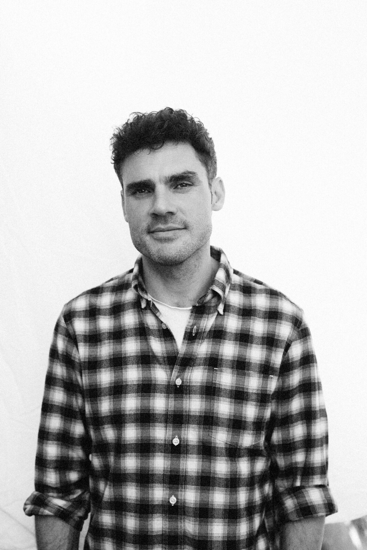 A man in a checkered shirt is standing in front of a simple background and looking at the camera. The picture is black and white.