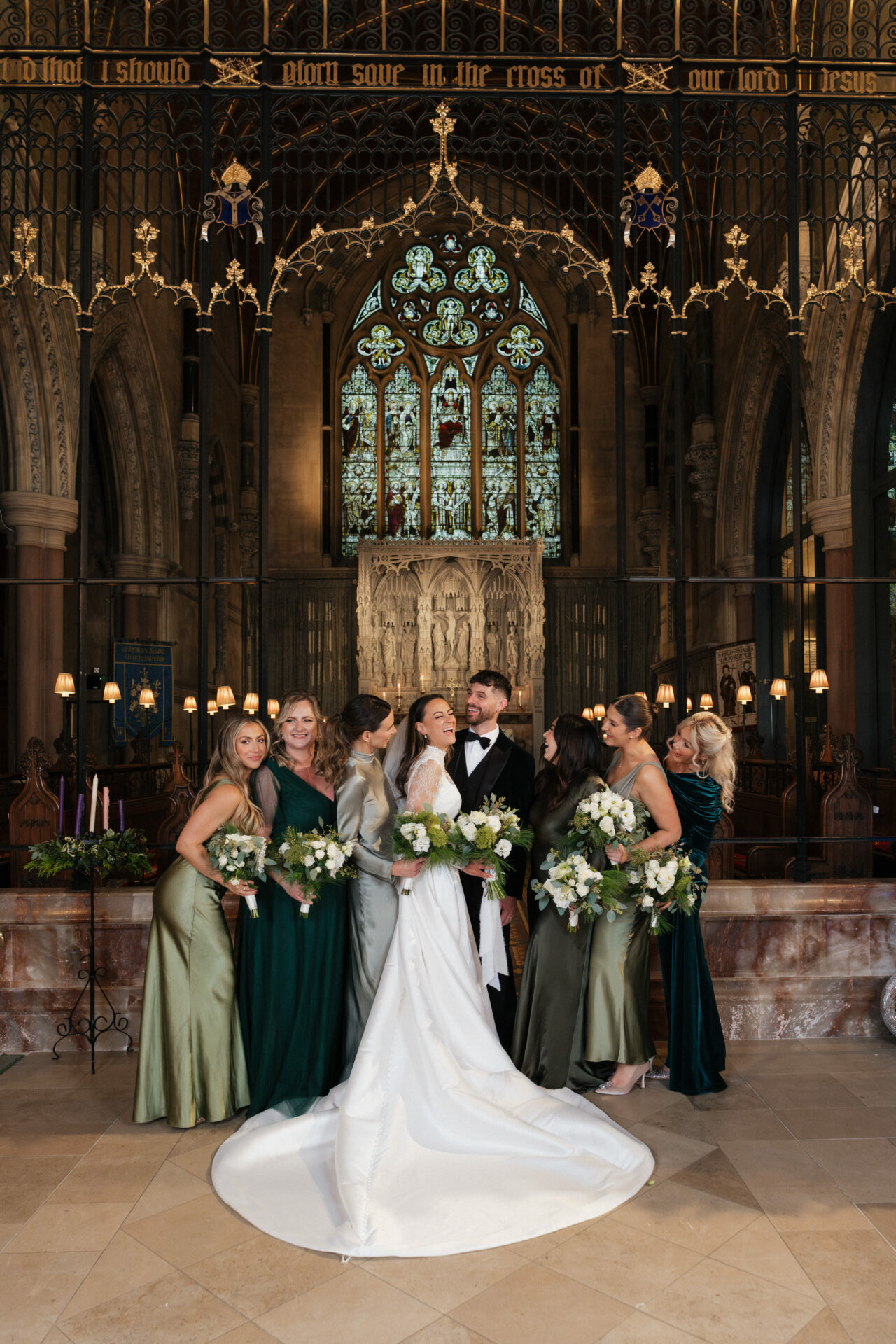 The bride and groom are with bridesmaids in green dresses holding flowers inside a decorated church with stained glass behind them.