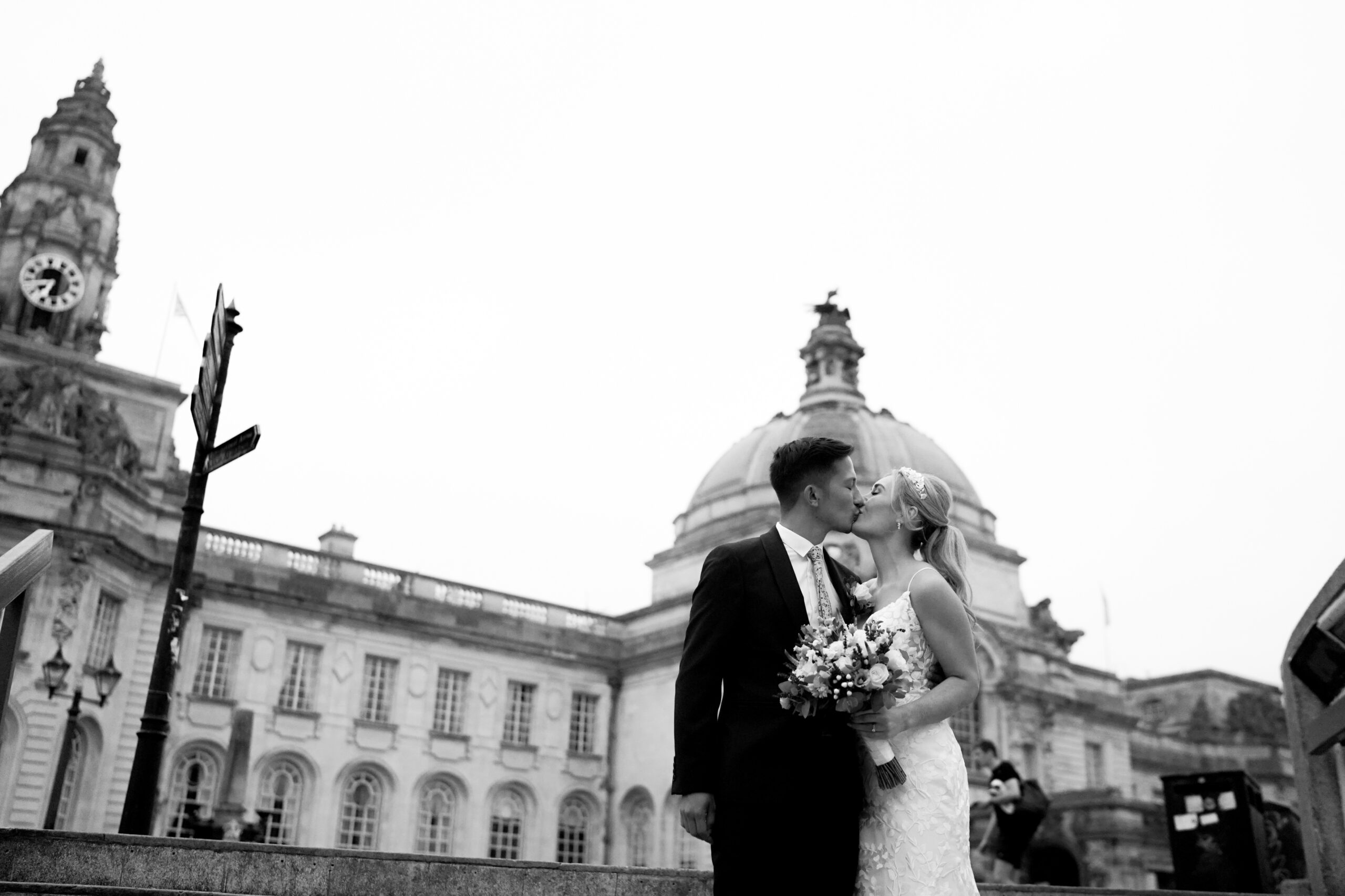 A bride and groom kiss in front of a fancy building with a dome and clock tower.