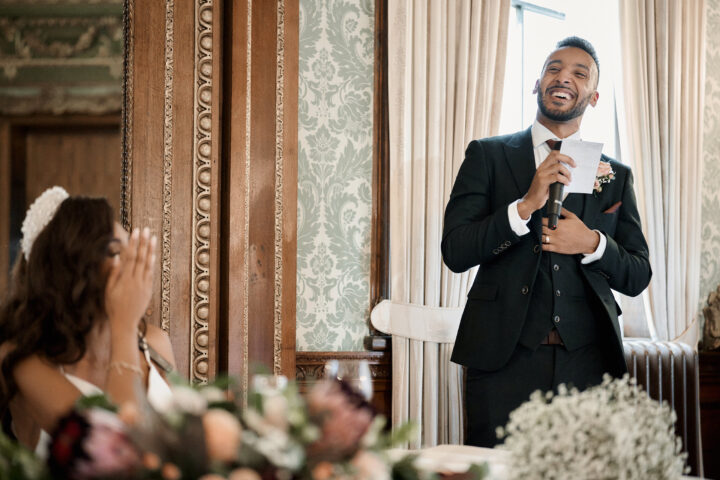 A man giving a speech and grinning at his wife, who's seated at a table in a fancy old-fashioned room.