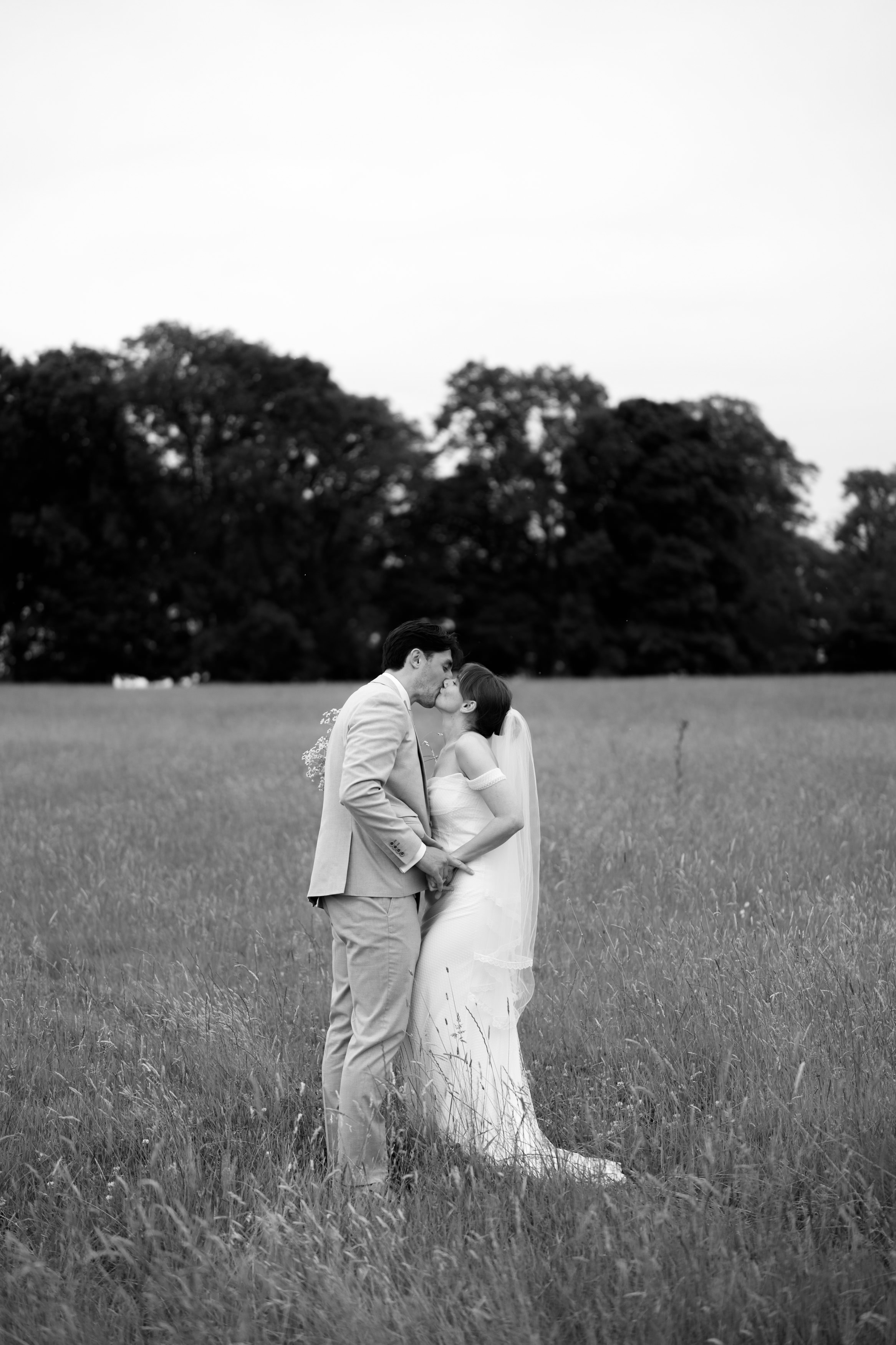 A couple in their wedding clothes kisses in a grassy field with trees behind them. The picture is in black and white.