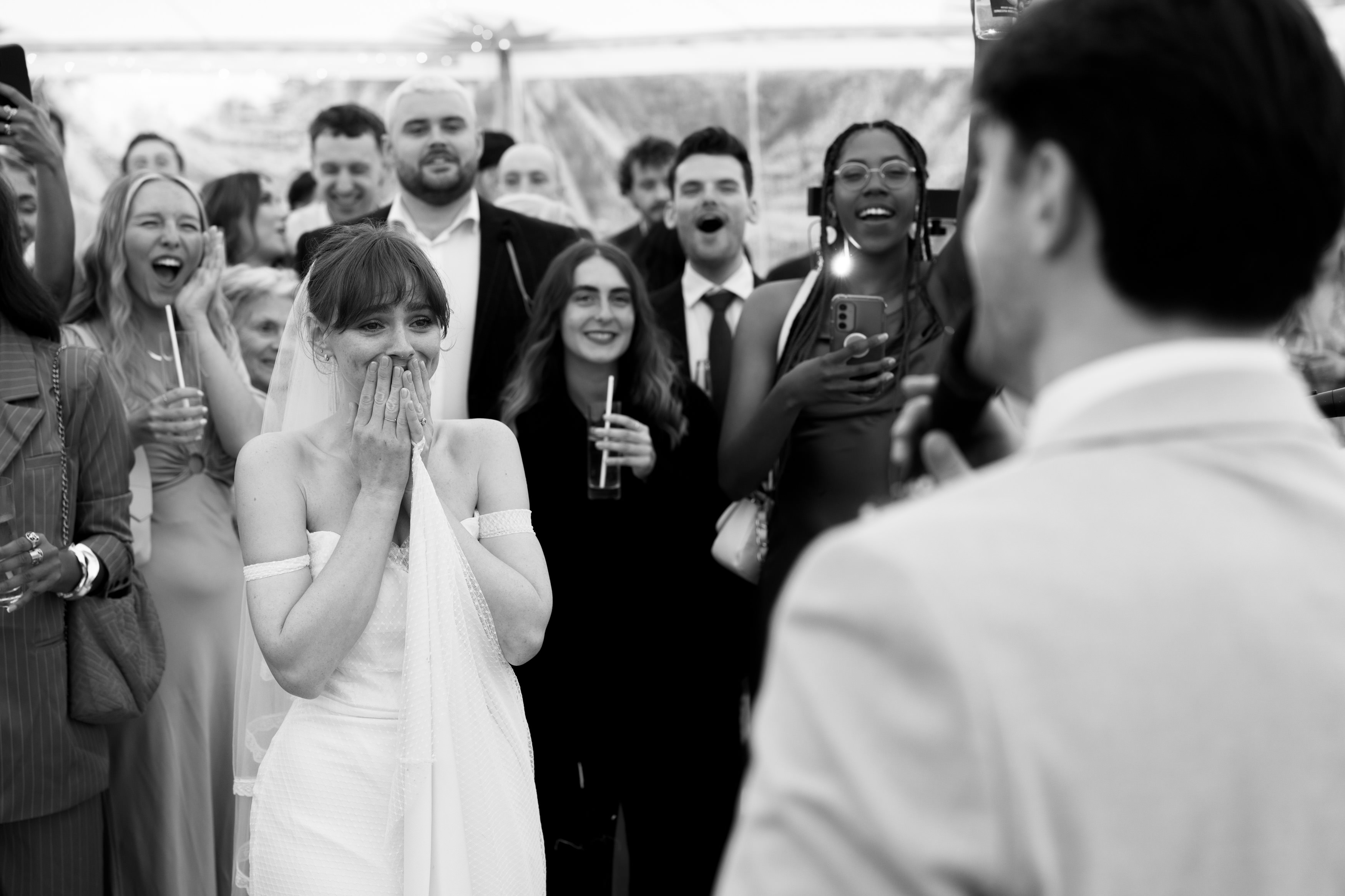 A bride in a white dress looks surprised and moved as she stands with smiling guests around her. A man in a suit is facing her at this lively get-together.