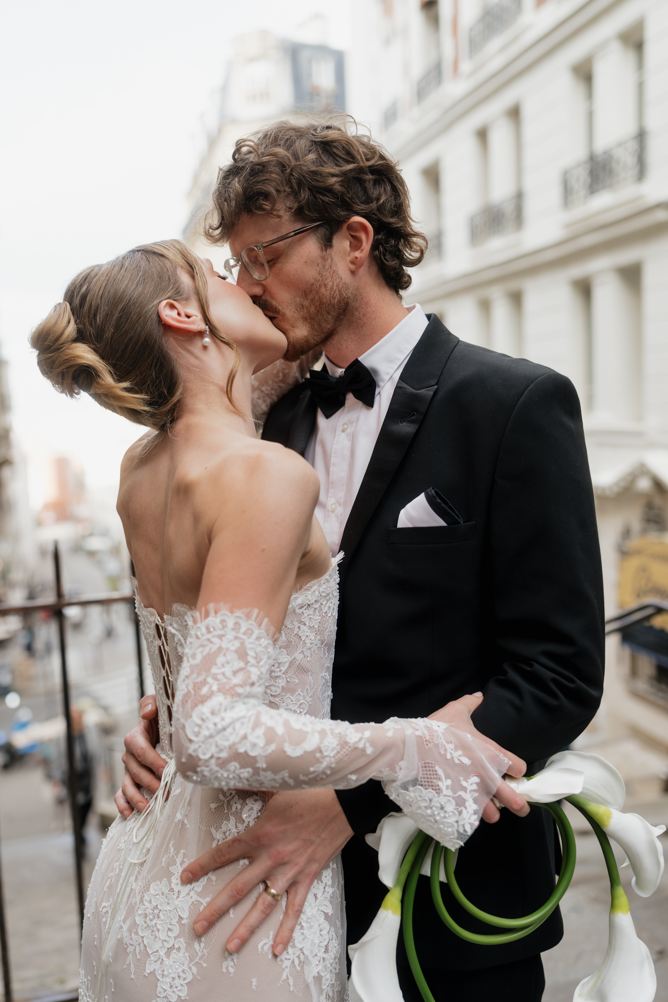 A bride and groom kiss on a city balcony. The bride is holding white calla lilies, and city buildings can be seen behind them.