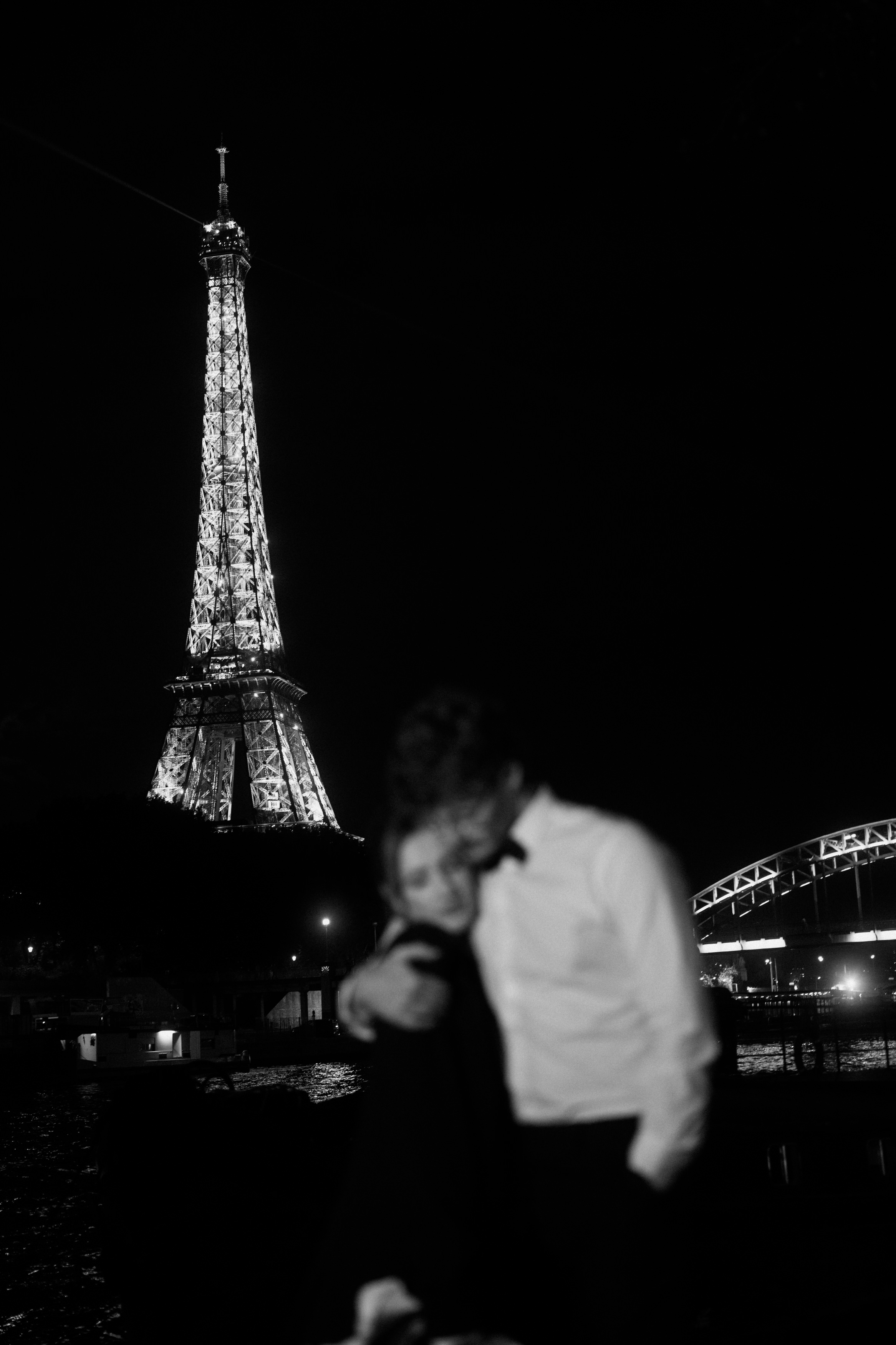 A couple stands out of focus at night, while the brightly lit Eiffel Tower clearly appears in the background.