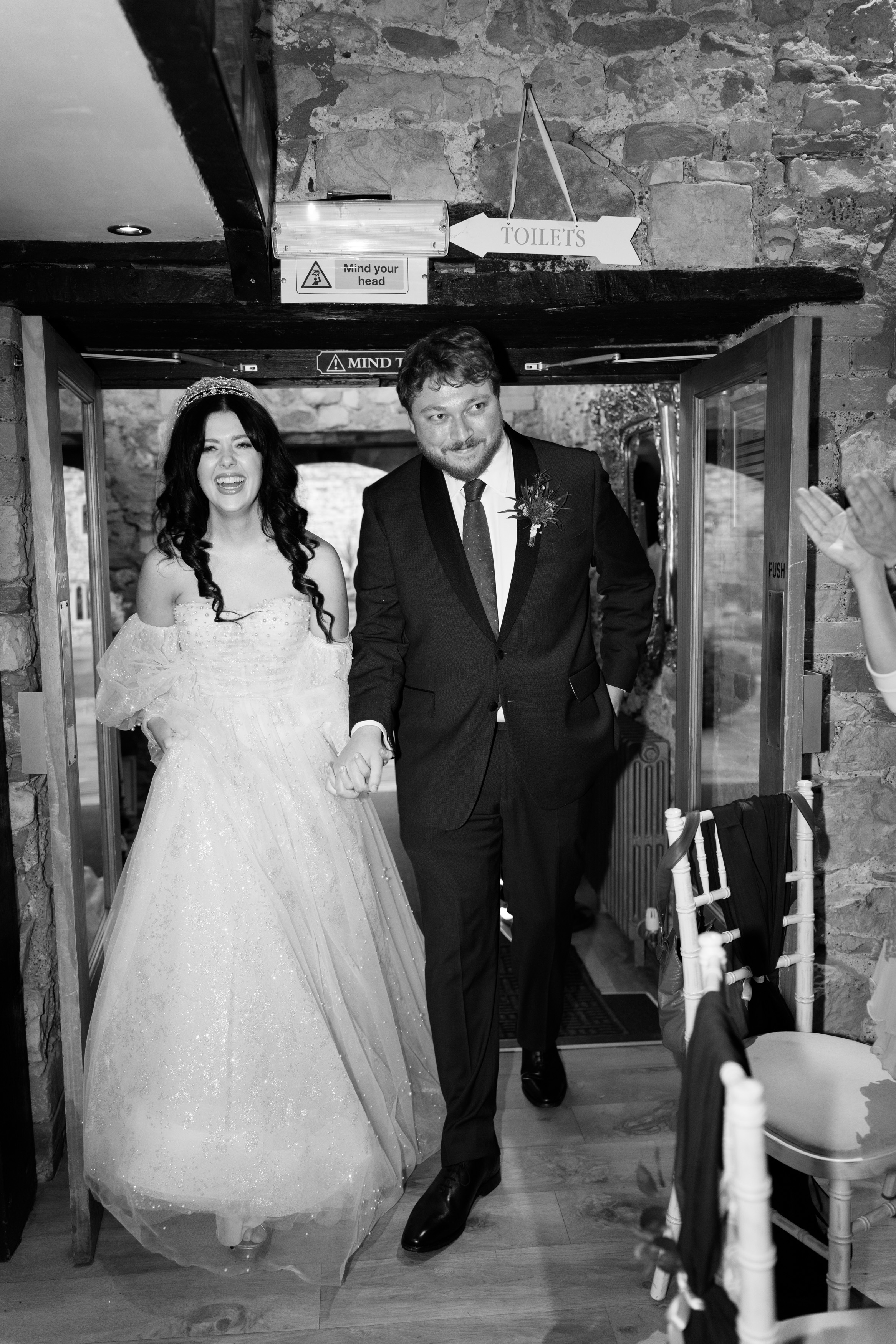 A bride and groom walk hand in hand through a doorway, smiling. The bride is wearing a white dress, and the groom has on a dark suit. There's a "Toilets" sign above them.