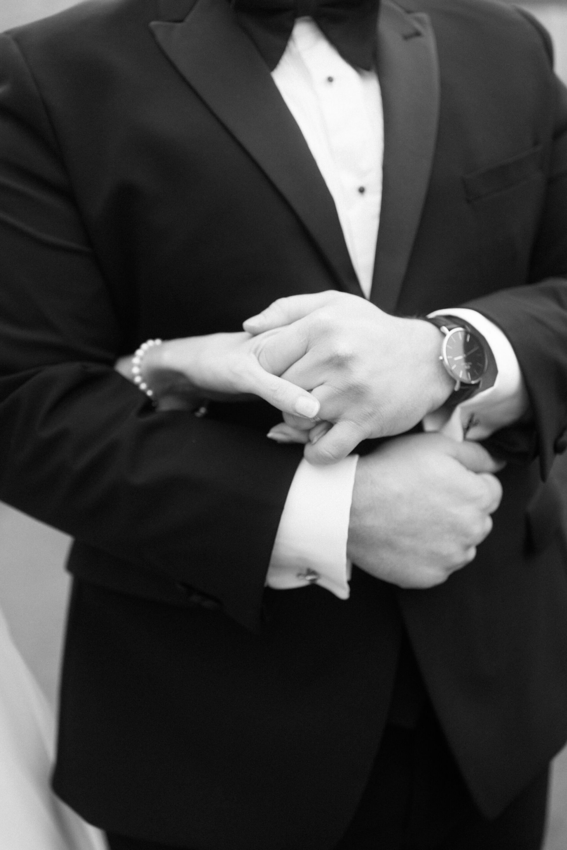 A couple is shown dressed up nicely. The man is wearing a suit with a bow tie and has his hands together, which shows off his watch. The woman is softly holding onto his arm, and you can see she's wearing a pearl bracelet on her wrist.