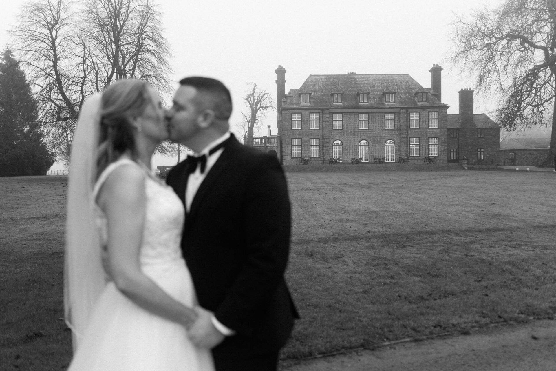 A bride and groom kiss outside a big, old building on a foggy day.