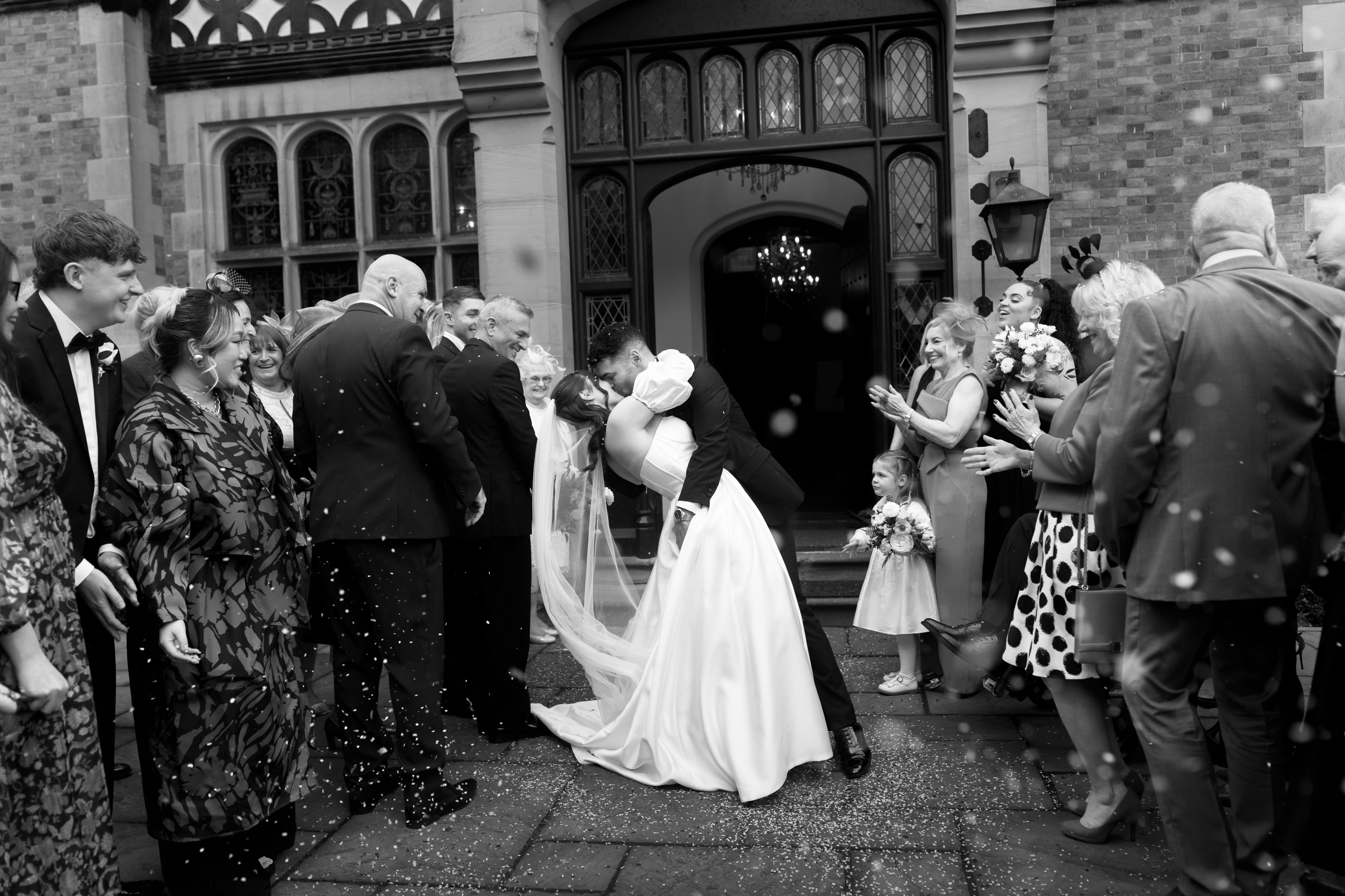 A couple kisses during their wedding outside a building, while guests cheer and flower petals float through the air.