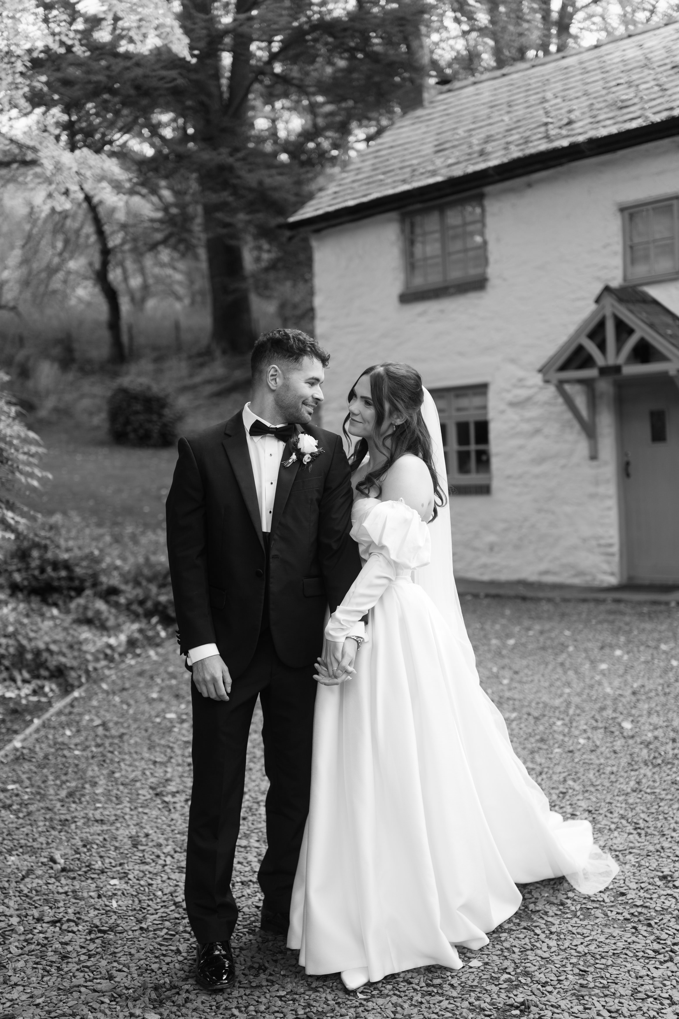 A couple dressed for their wedding is standing outside a charming, old-fashioned house. The groom is wearing a suit with a bow tie, and the bride is in a long dress with a veil. They are smiling at each other.