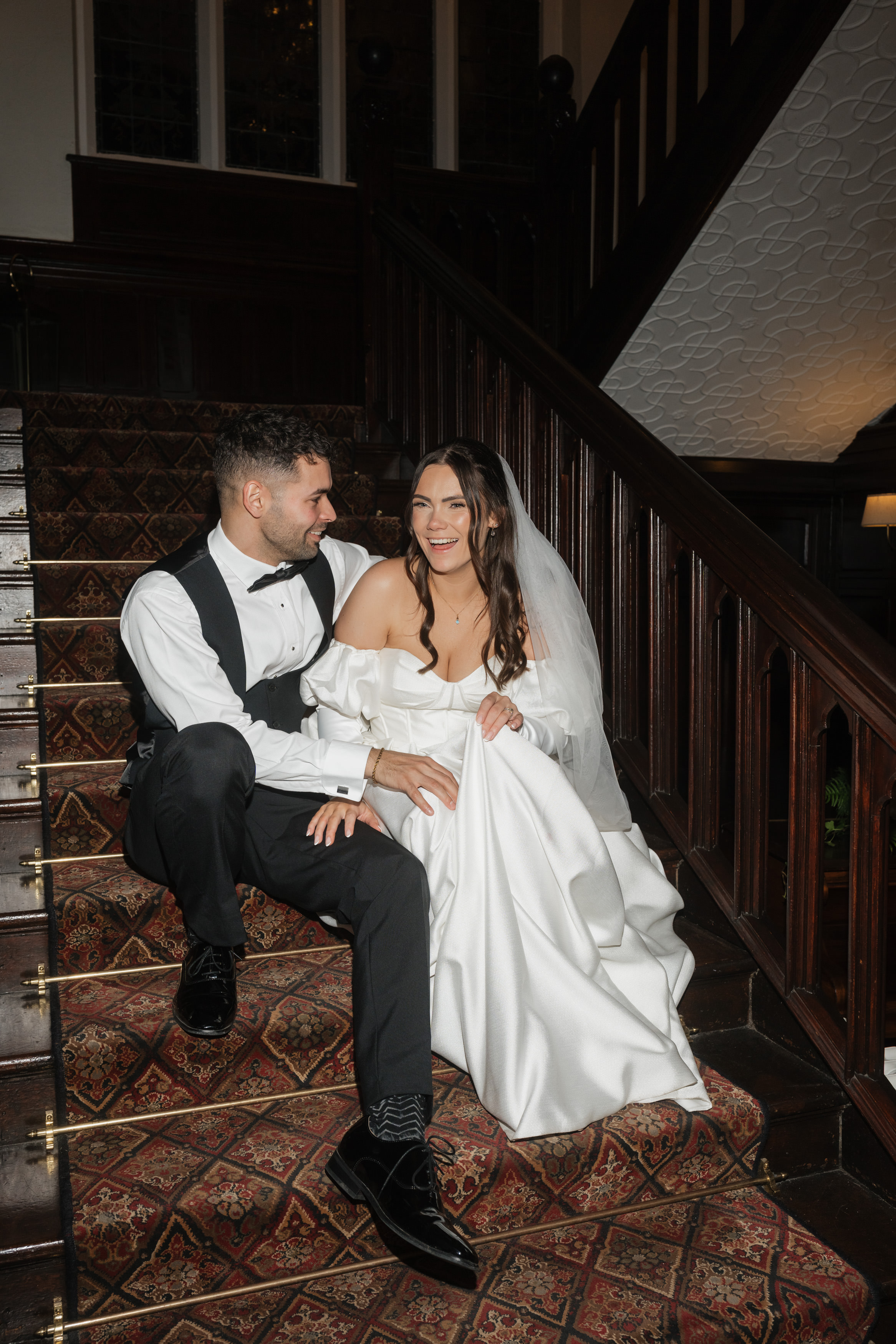 A bride and groom are sitting on a carpeted staircase, happily smiling at each other. The bride is wearing an off-the-shoulder dress with a veil, and the groom is dressed in a tuxedo.