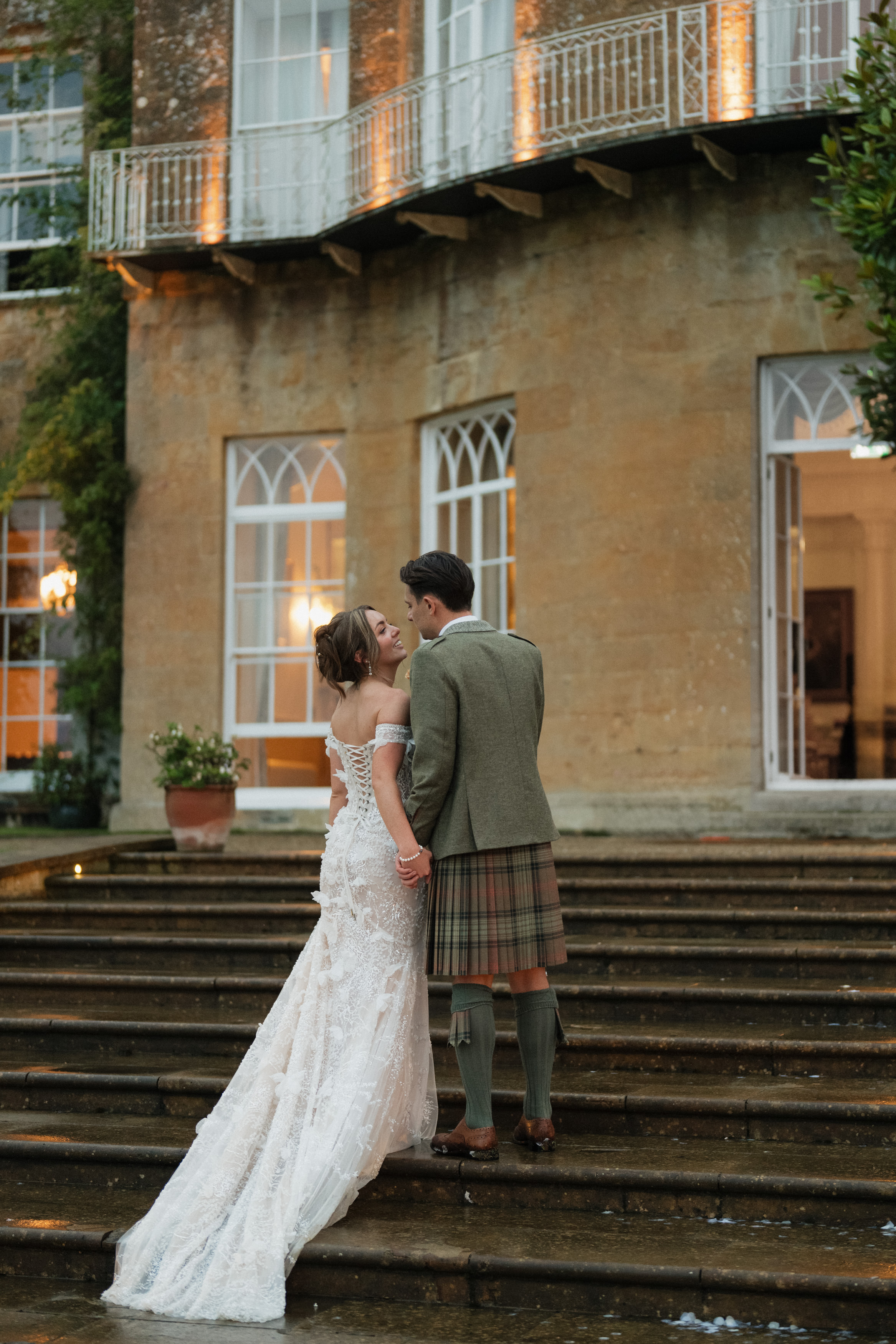 A couple is holding hands on the steps of an old building. The woman is wearing a white dress, and the man is dressed in a kilt and a jacket.