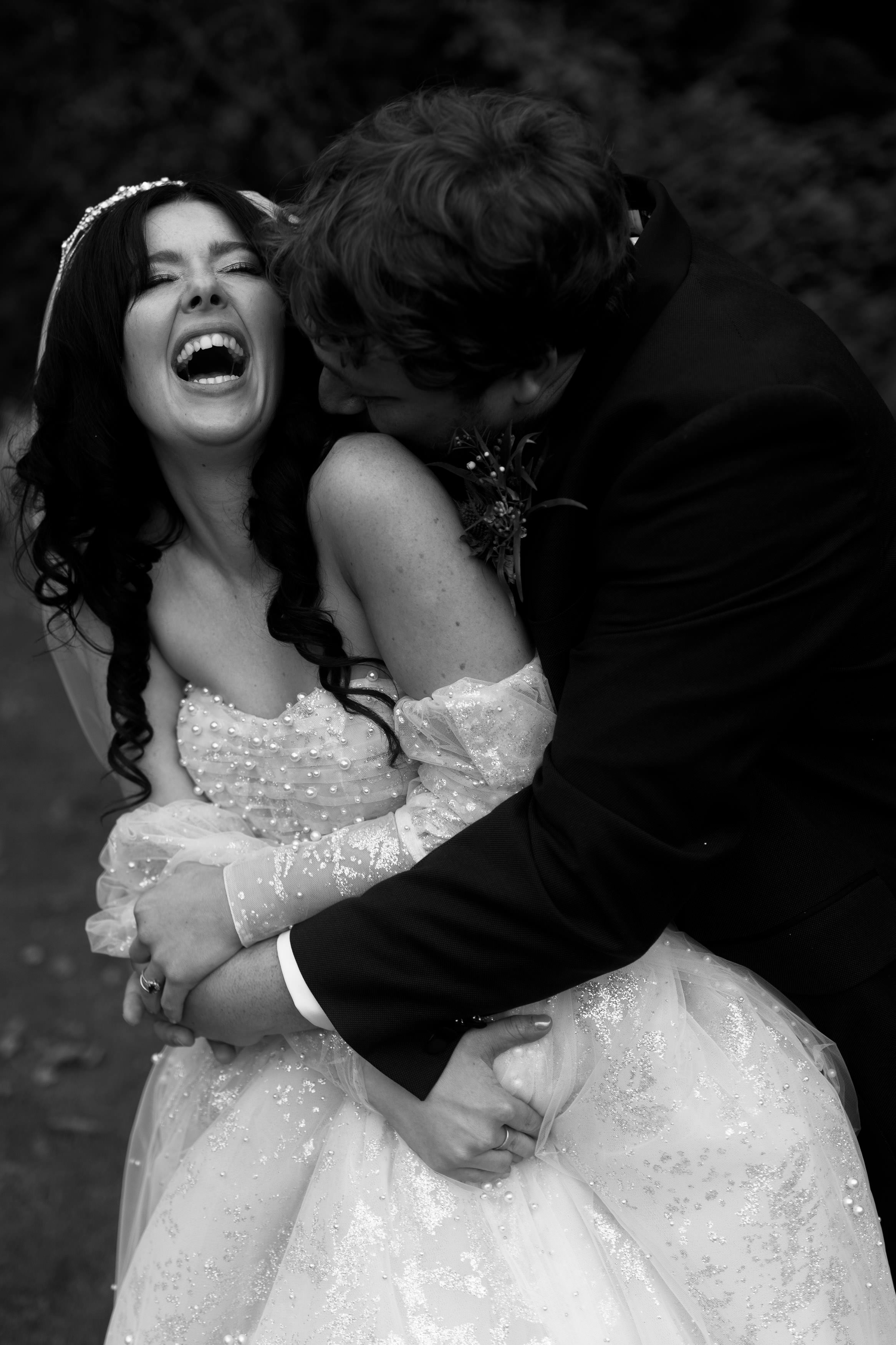 A couple dressed in wedding clothes shares a happy moment together. The bride is laughing while the groom gives her a hug from behind.