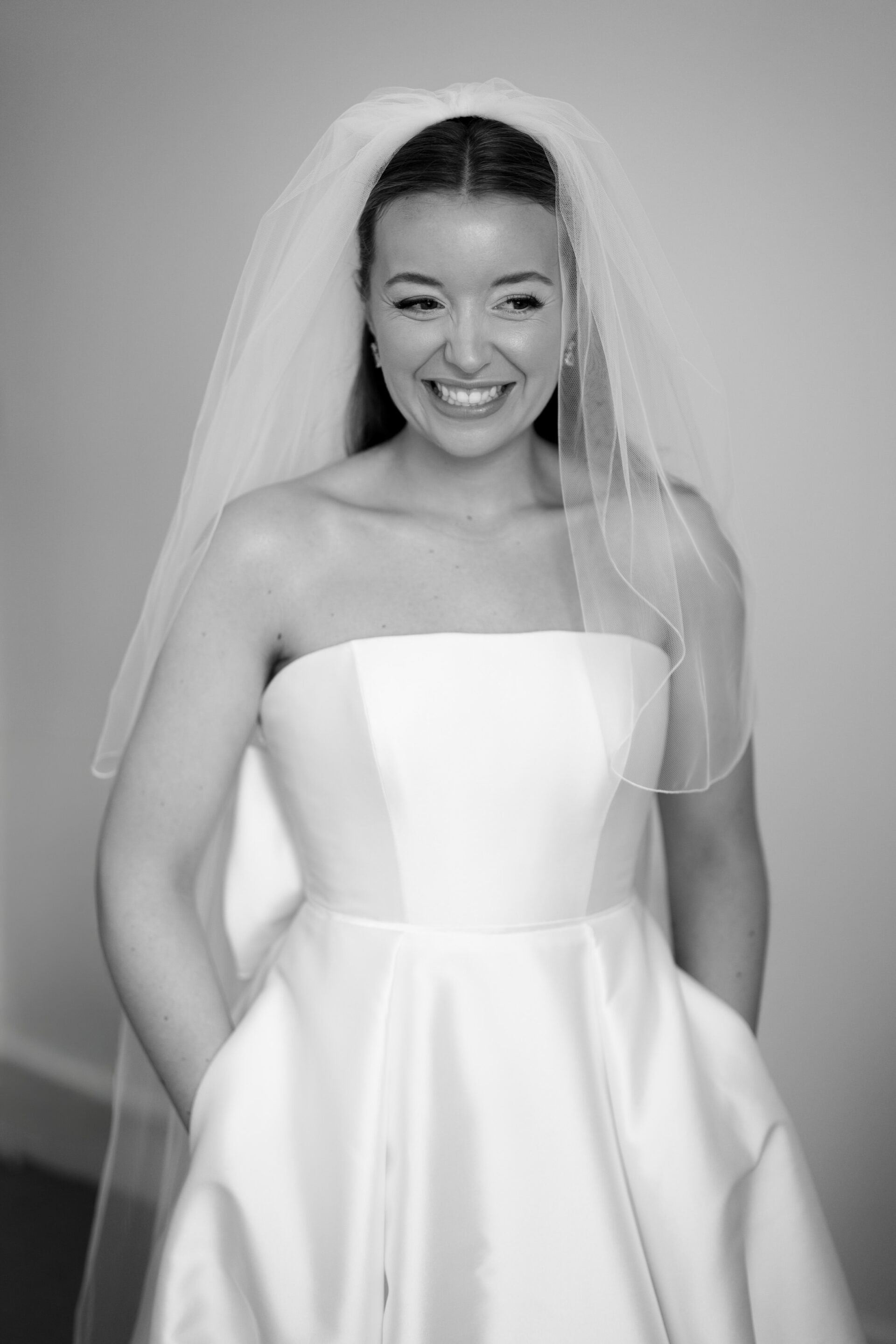 A bride in a white sleeveless dress and veil is smiling as she stands inside.