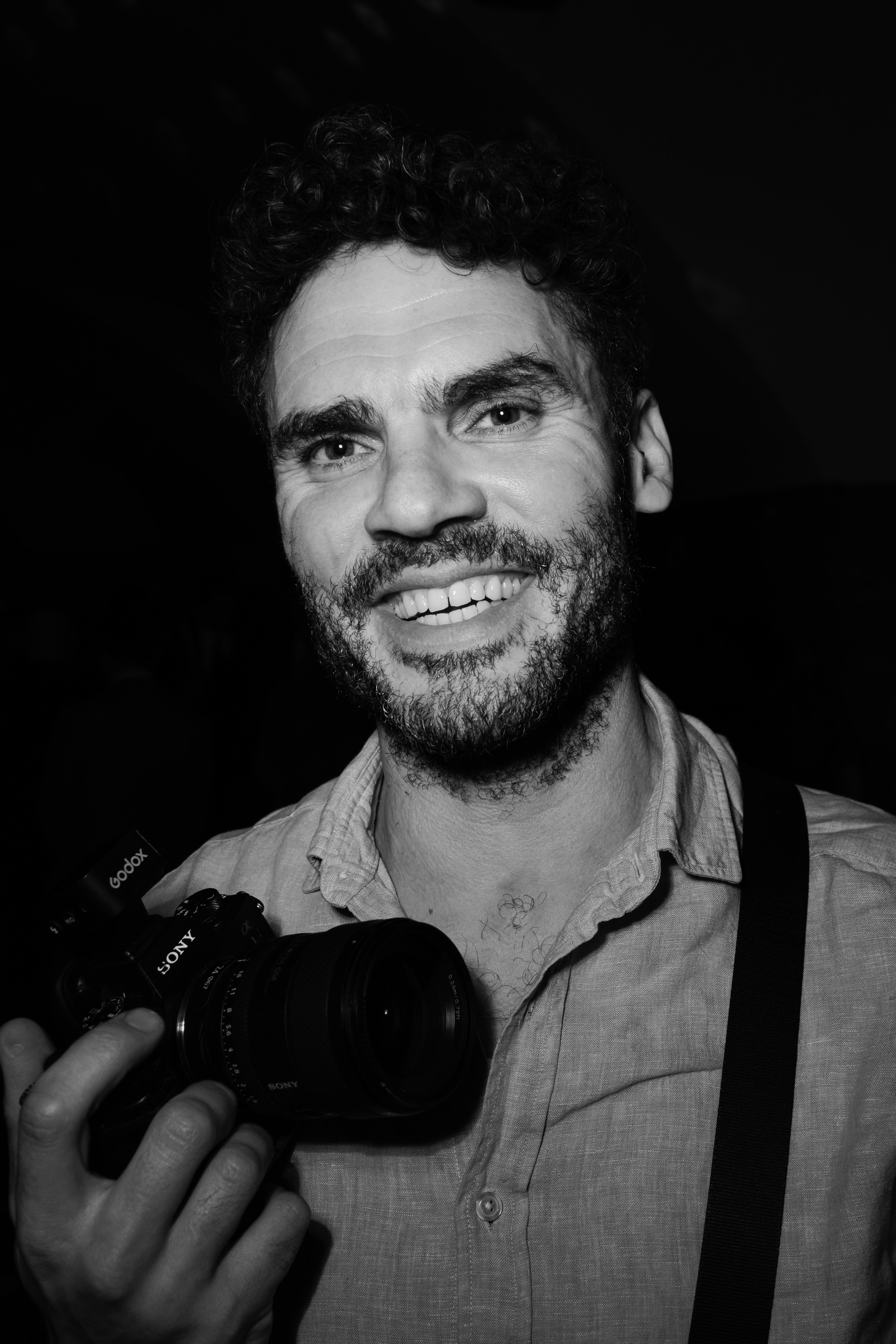 A smiling man with curly hair and a beard holds a Sony camera. He’s wearing a button-up shirt and has a strap over his shoulder.