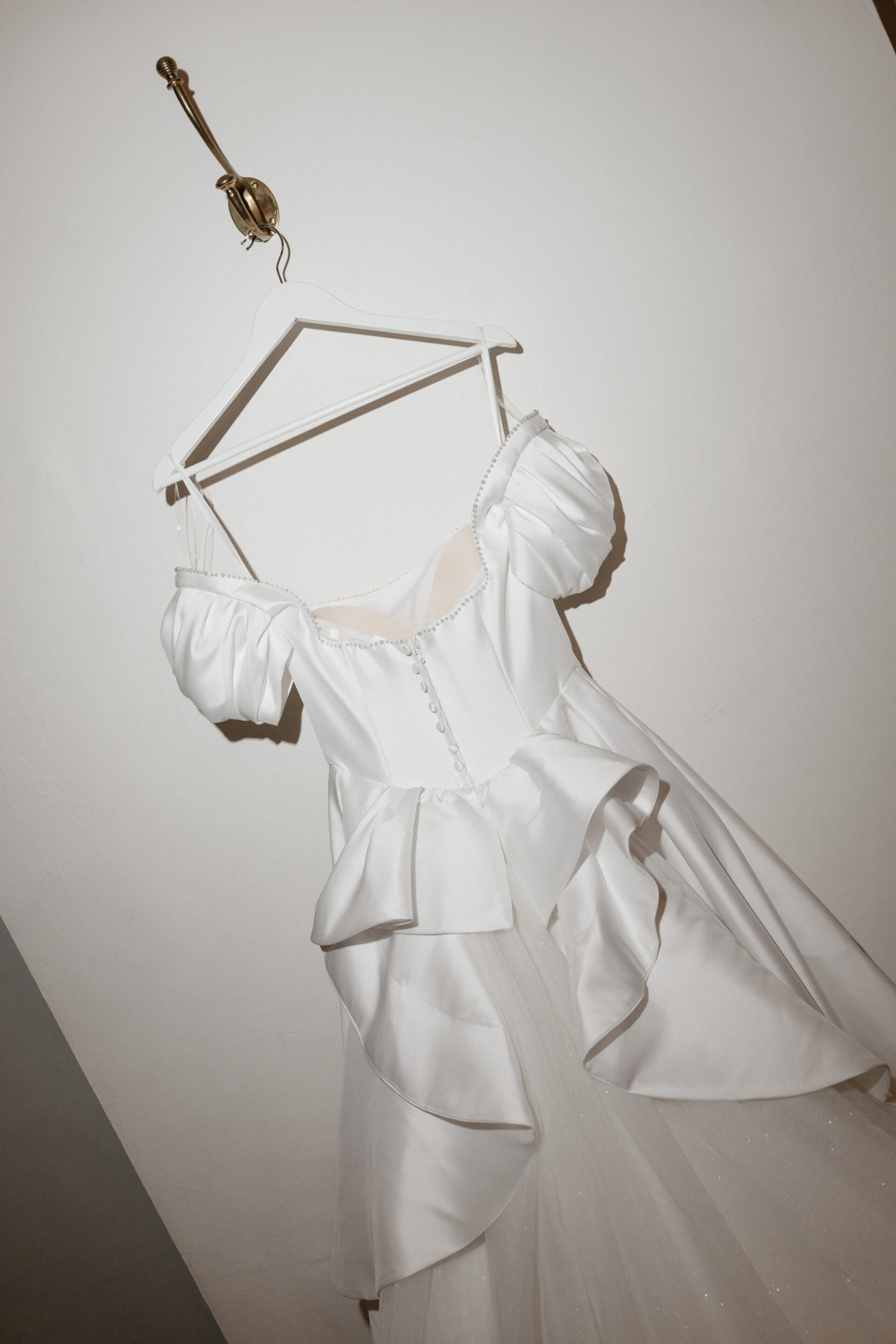 A white wedding dress with puffy sleeves and a layered skirt hangs on a hanger from a hook on a white wall.