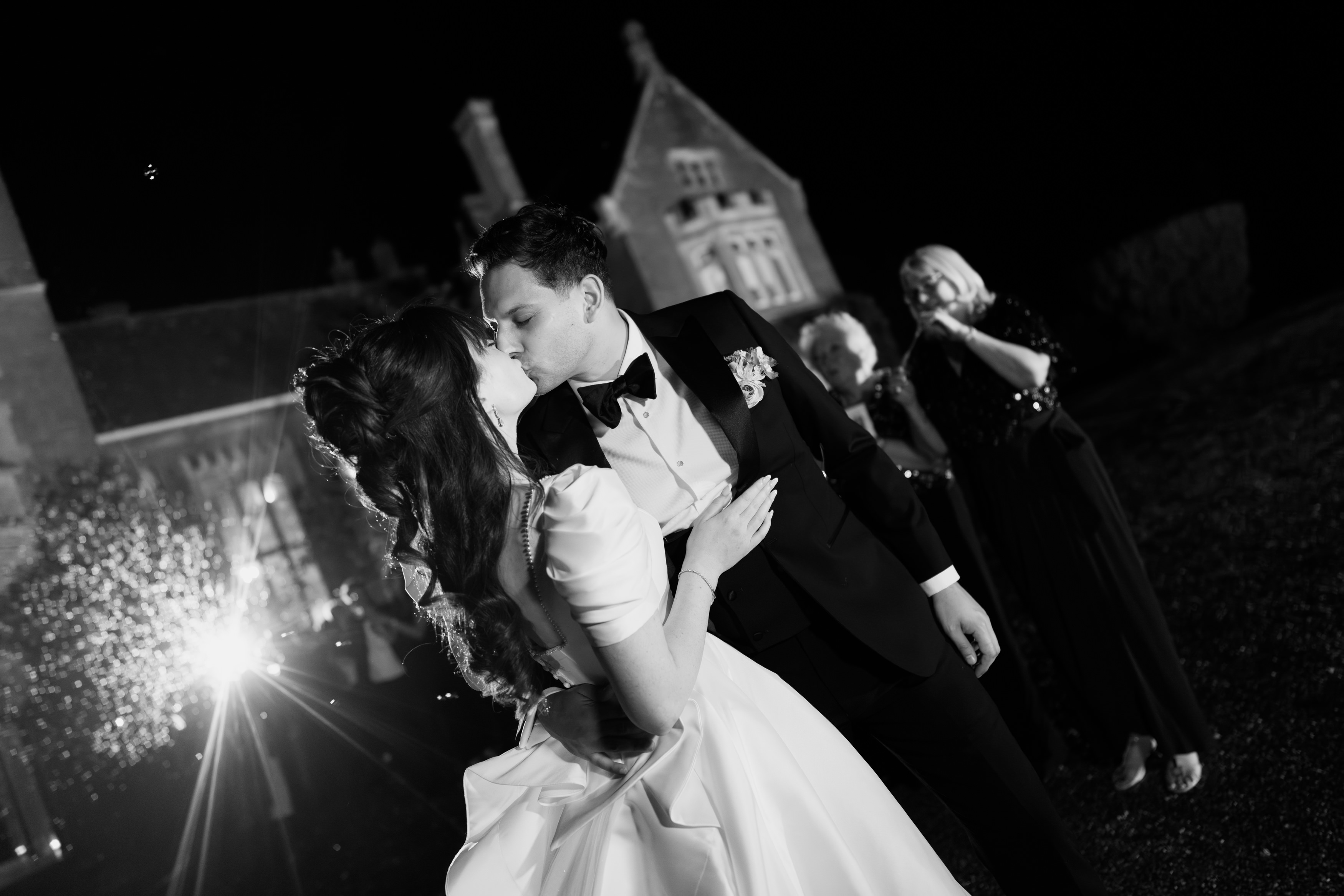 A bride and groom kiss outside at night by a big building, wearing wedding clothes, with guests seen in the background.