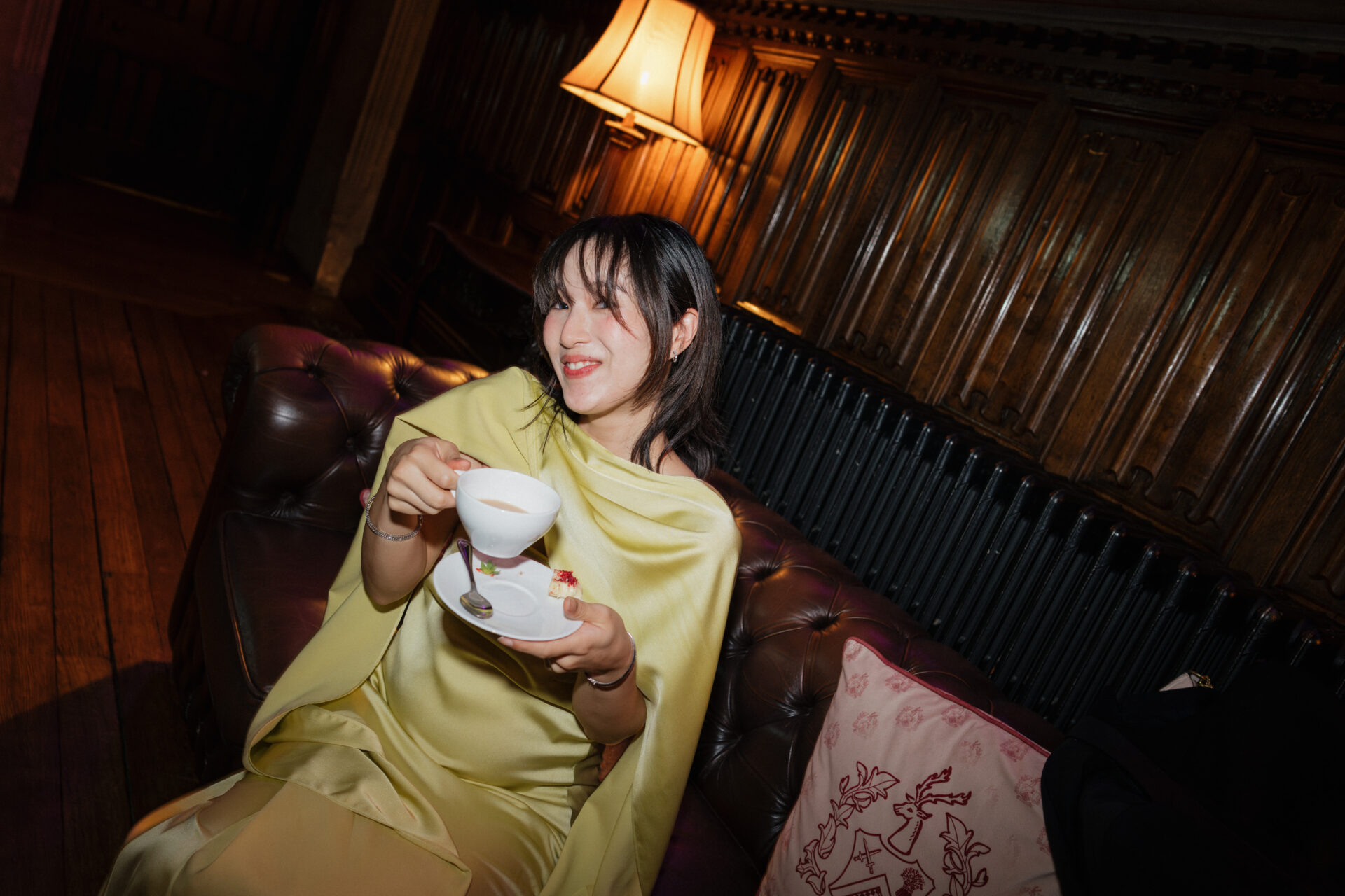 A woman in a bright yellow dress sits on a leather couch, smiling as she holds a teacup, with wood walls and a lamp behind her.