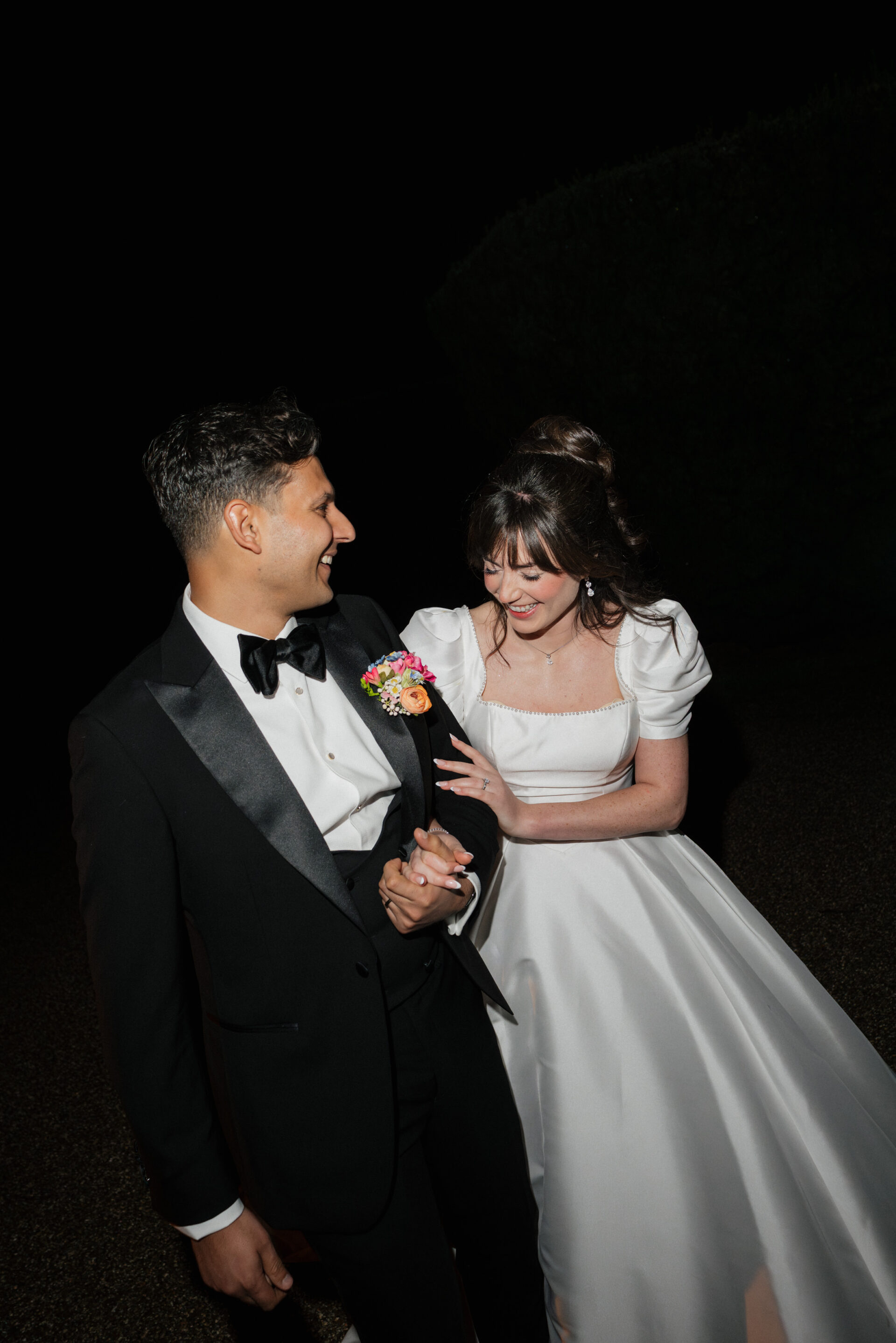 A bride in a white dress and a groom in a black tux hold hands and smile as they walk outside together at night.