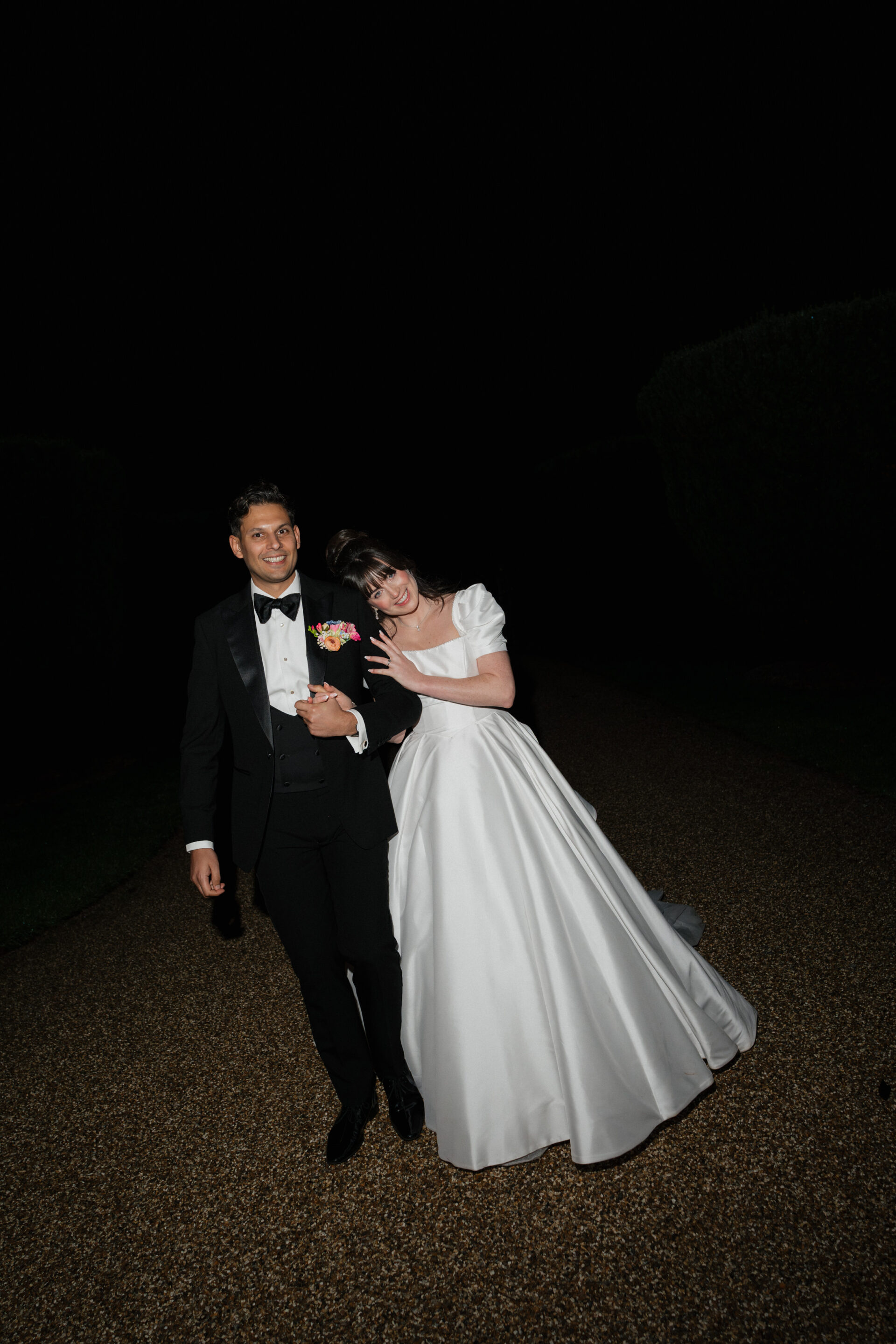 A bride in a white dress waves at the camera while walking outside at night with her groom, who’s in a black tux and bow tie.