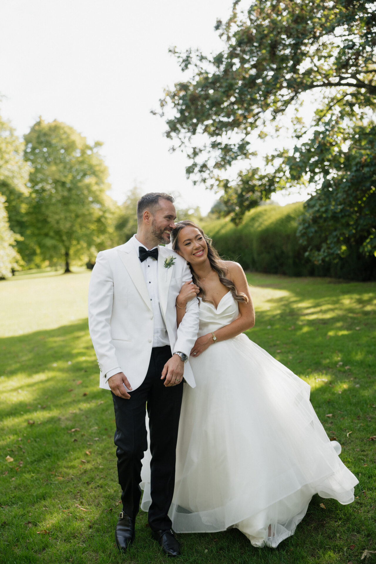 A bride in a white dress and a groom in a white jacket and black pants smile together on the grass with trees behind them.
