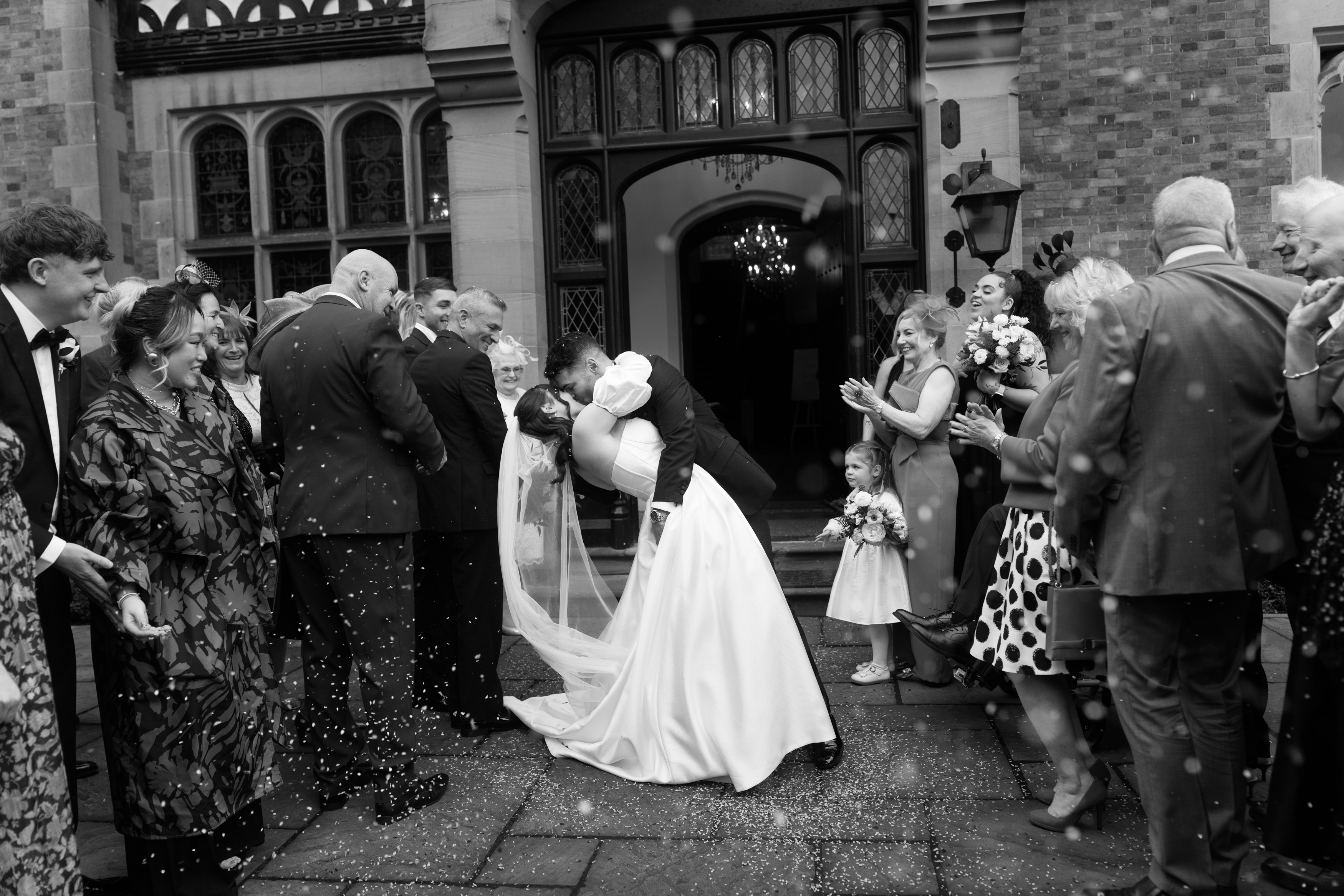 A bride and groom share a passionate kiss outside a building while guests gather around them. Confetti is tossed in the air as part of the celebration.