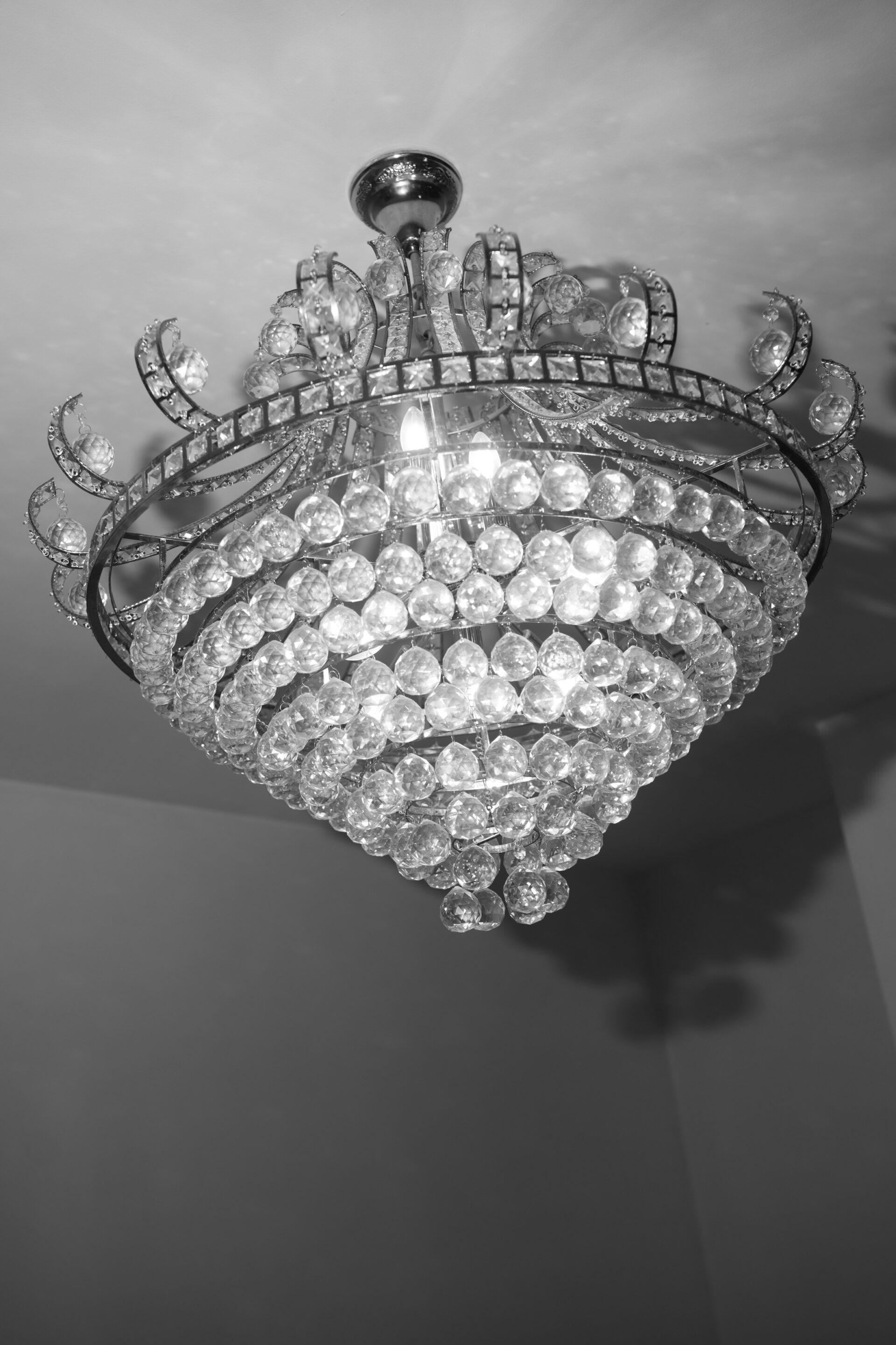 A black and white photo of a fancy crystal chandelier with a round shape. It's hanging from the ceiling and creating soft shadows.