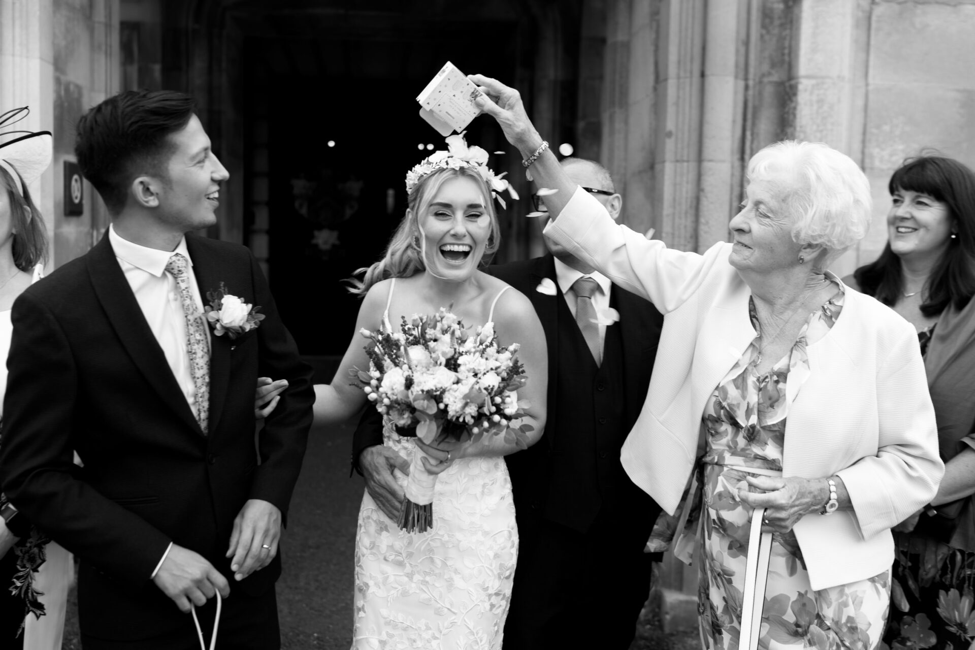 A happy couple is leaving a building, smiling, while an older woman playfully throws confetti over them.