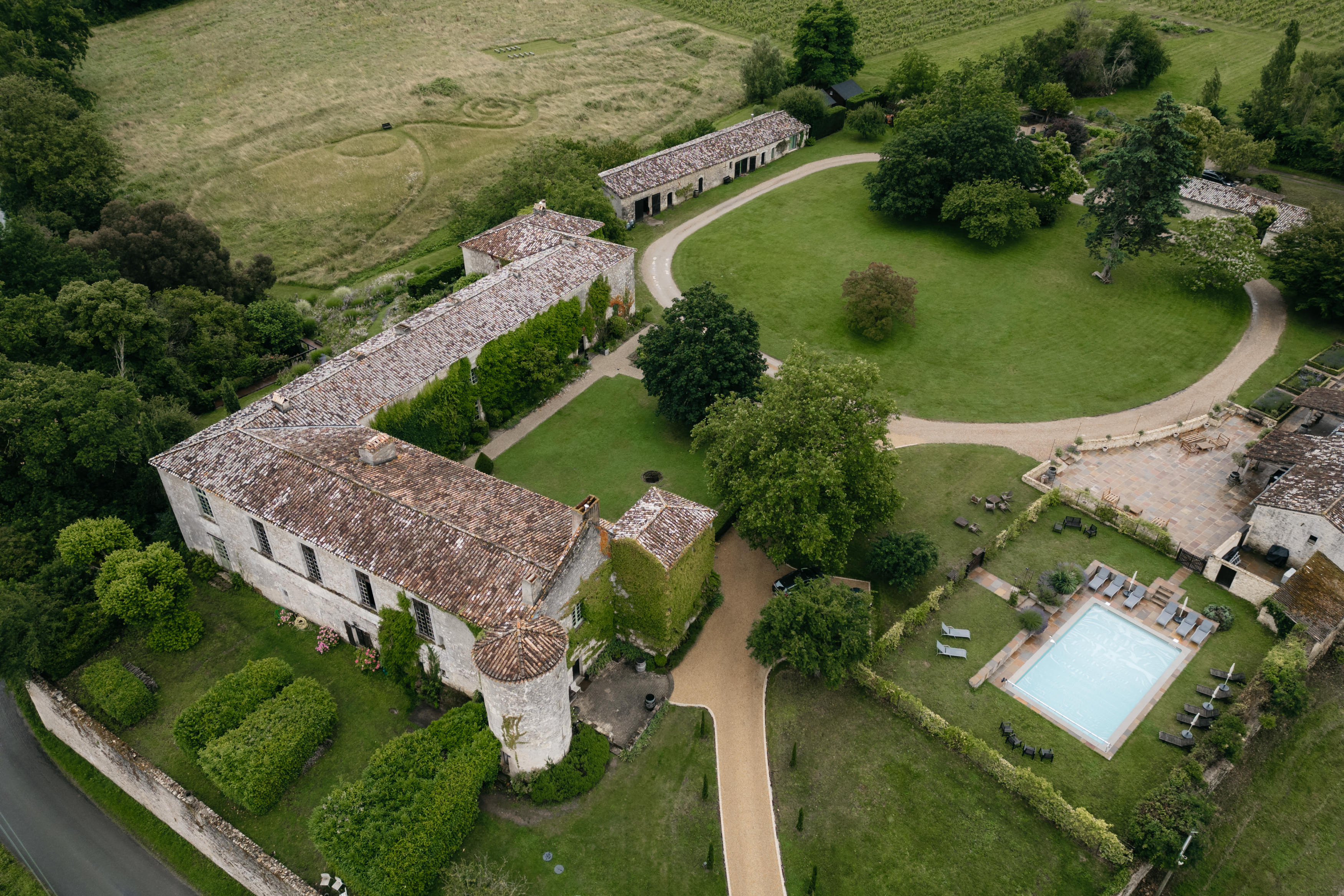 A view from above showing an old stone building with a red roof. It's surrounded by gardens, walkways, and a swimming pool.