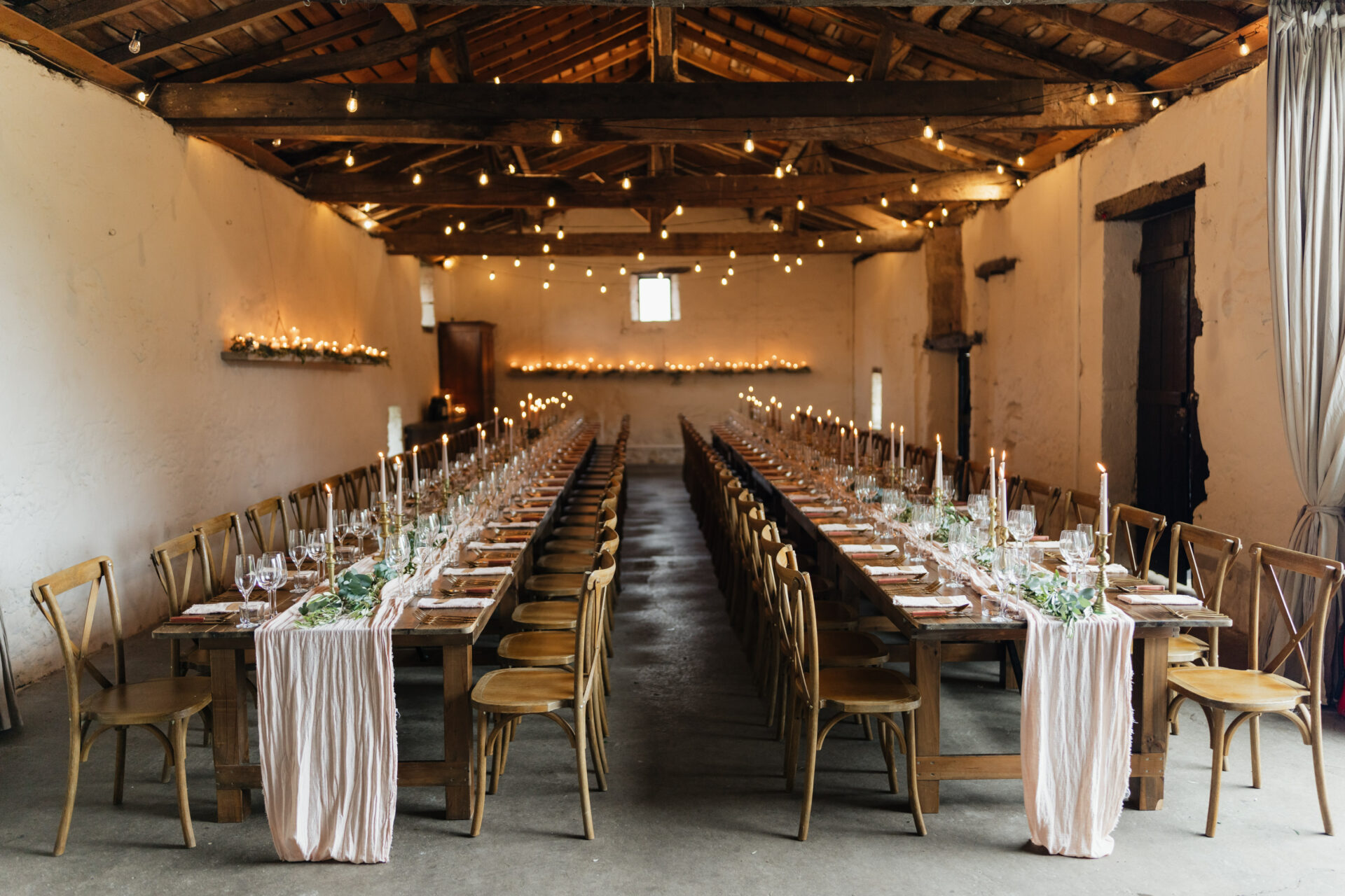 A cozy banquet hall with long wooden tables covered in pink tablecloths, ready for a fancy event. Soft lighting and candles make the place feel warm and inviting.