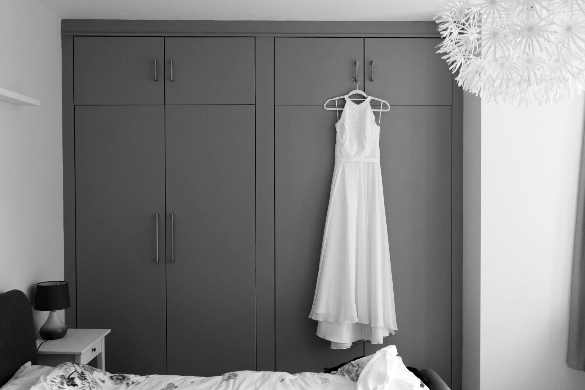 A sleeveless white dress is hanging on a gray closet door in a bedroom. The room also has a bed, nightstand, lamp, and a fancy ceiling light.