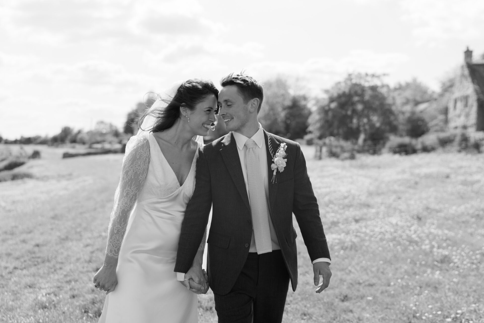 A bride and groom walk together, holding hands in a sunny field, smiling at one another. The bride is dressed in a lace-sleeved gown, while the groom wears a suit with a small flower pinned on it.