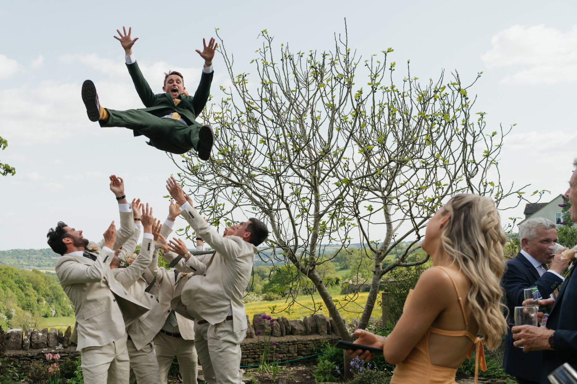 A group of people outside are throwing a man wearing a green suit into the air, with trees and fields all around them.